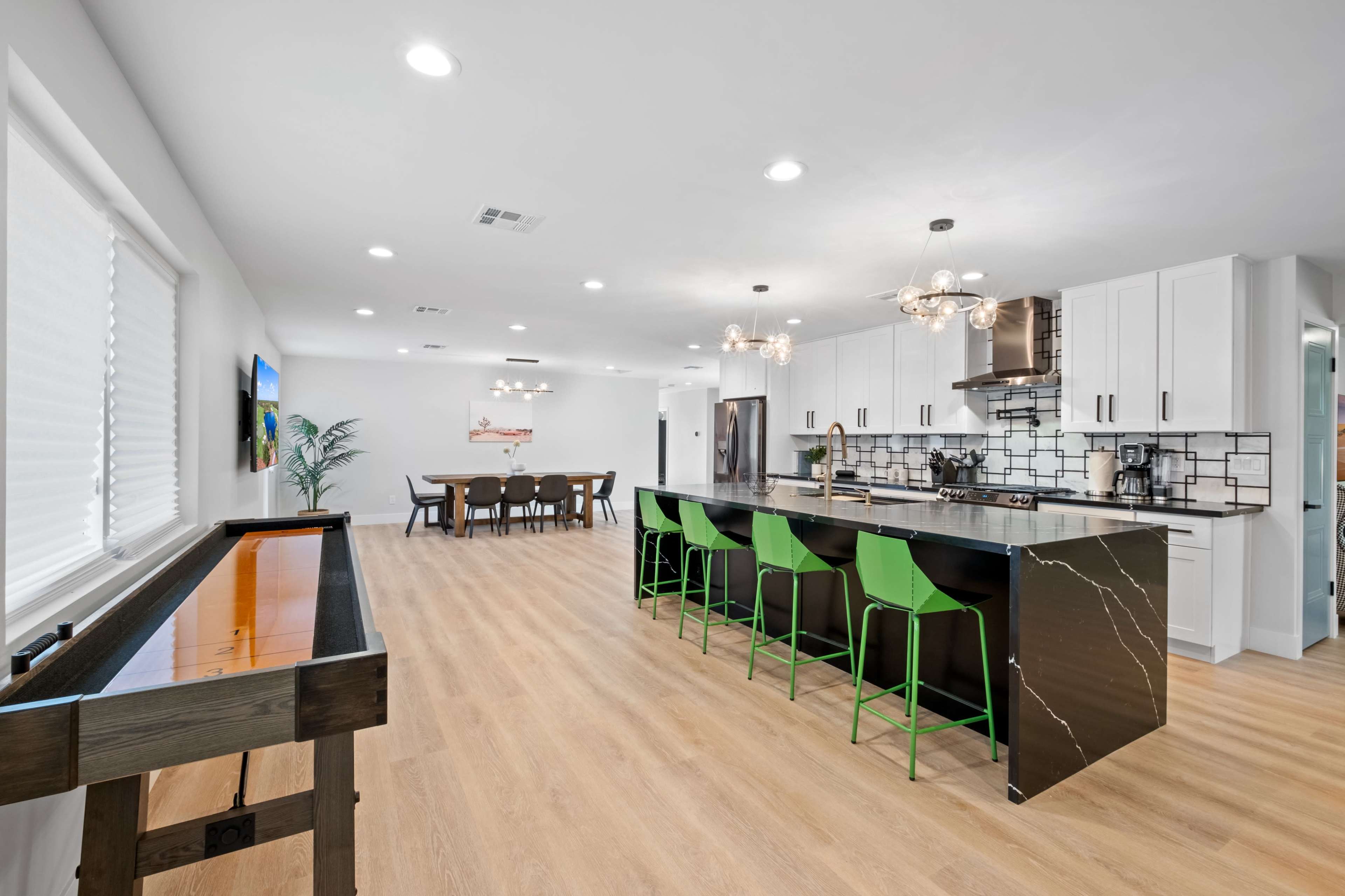 A modern kitchen with a long island featuring green stools, a dining area in the background, and light wooden flooring.