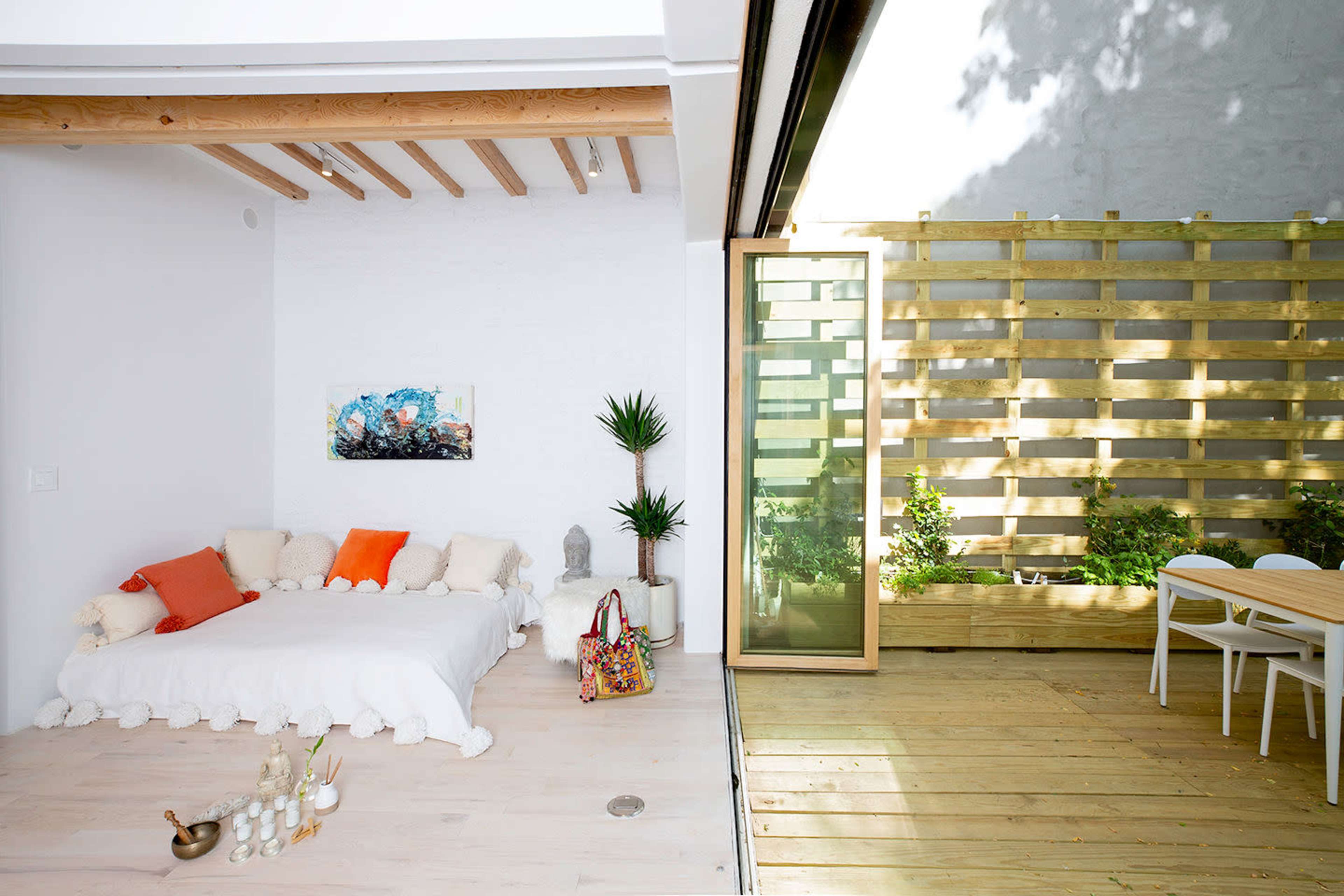 A bright, modern living space with a bed covered in light bedding on one side and a wooden deck with plants and a small dining area on the other.