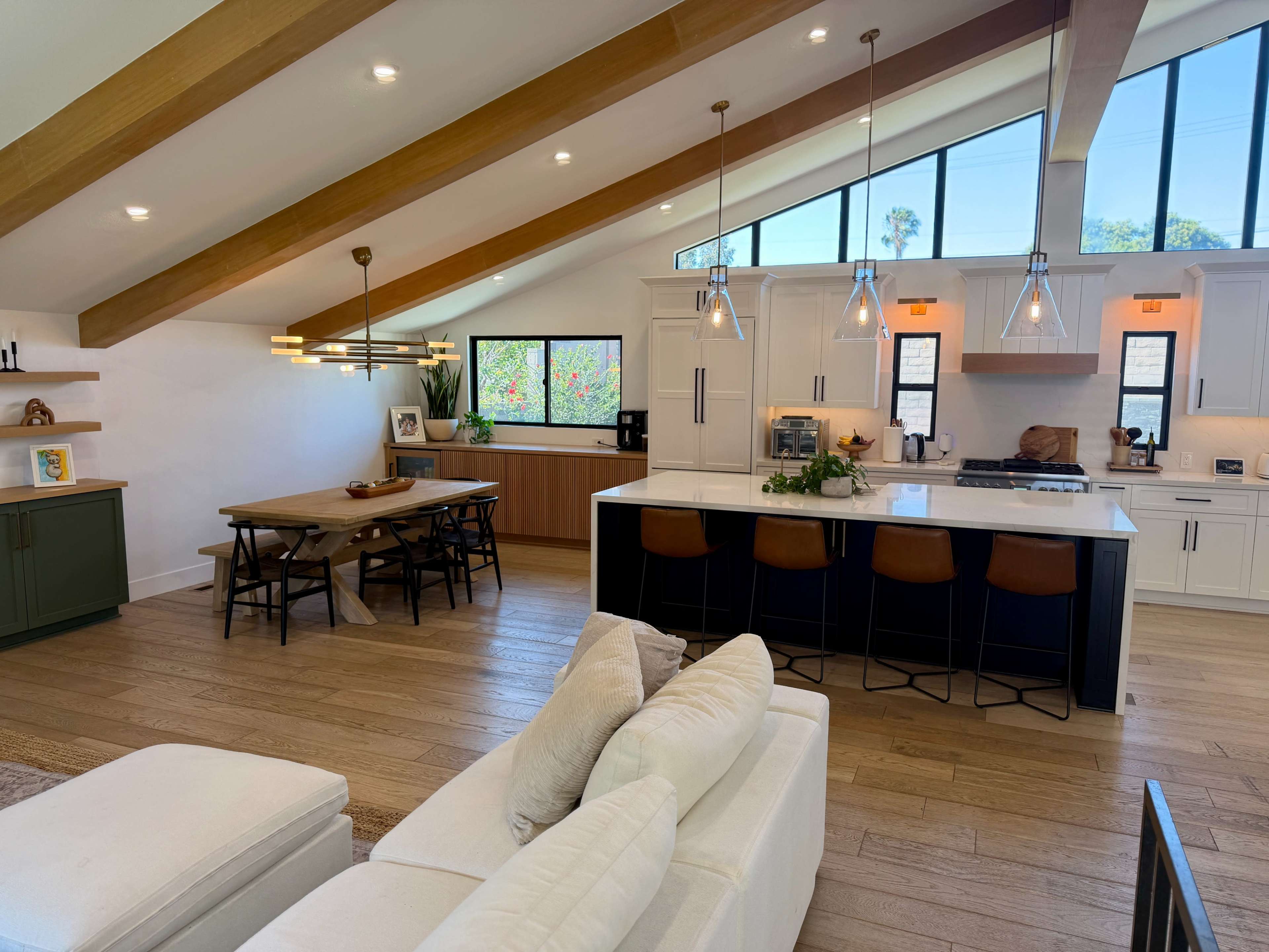 The image shows a modern open-concept living area with a dining table, kitchen bar stools, and large windows that provide natural light.