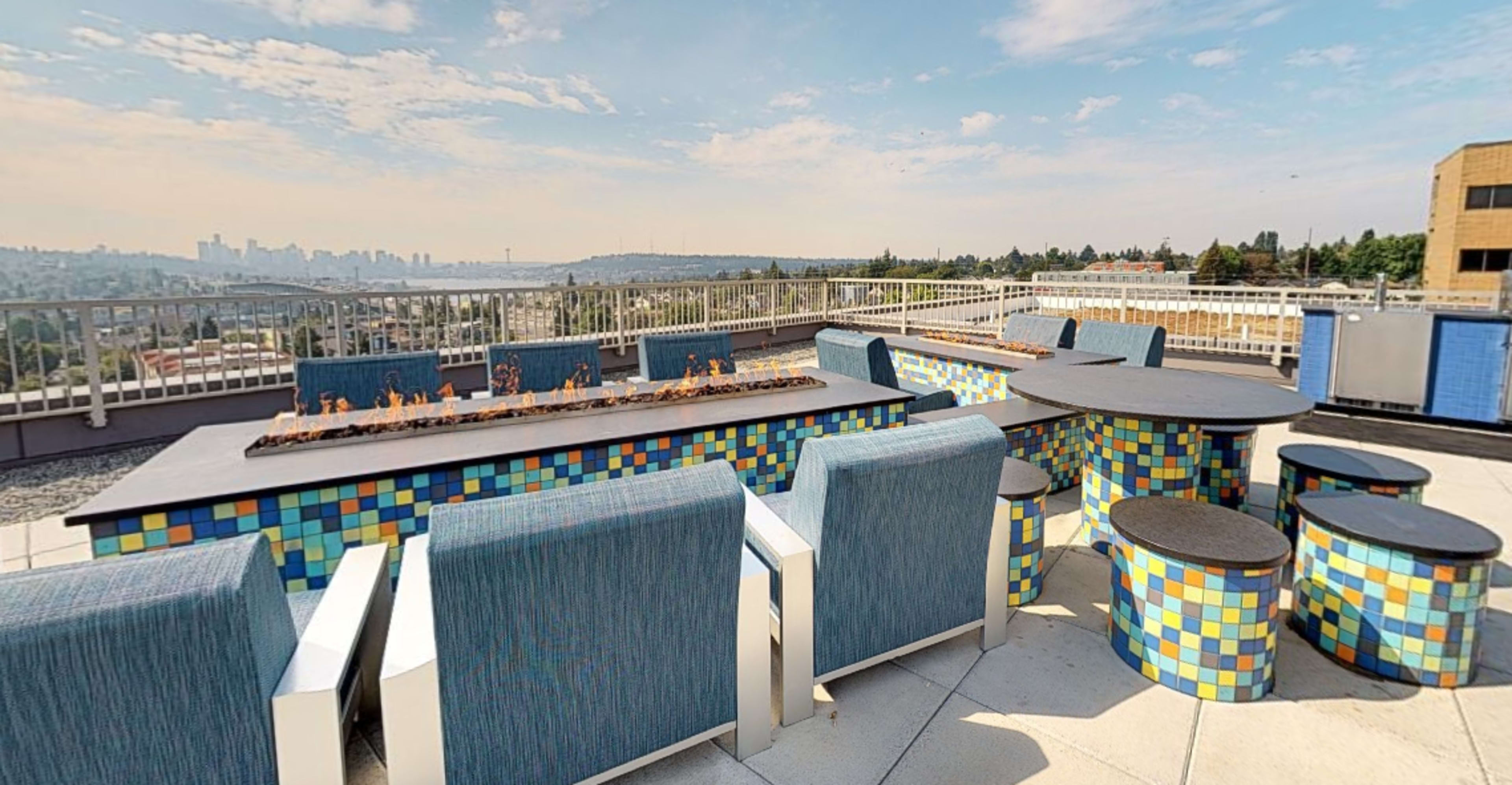 A rooftop patio with colorful tile furniture, including chairs and tables, overlooking a city skyline.