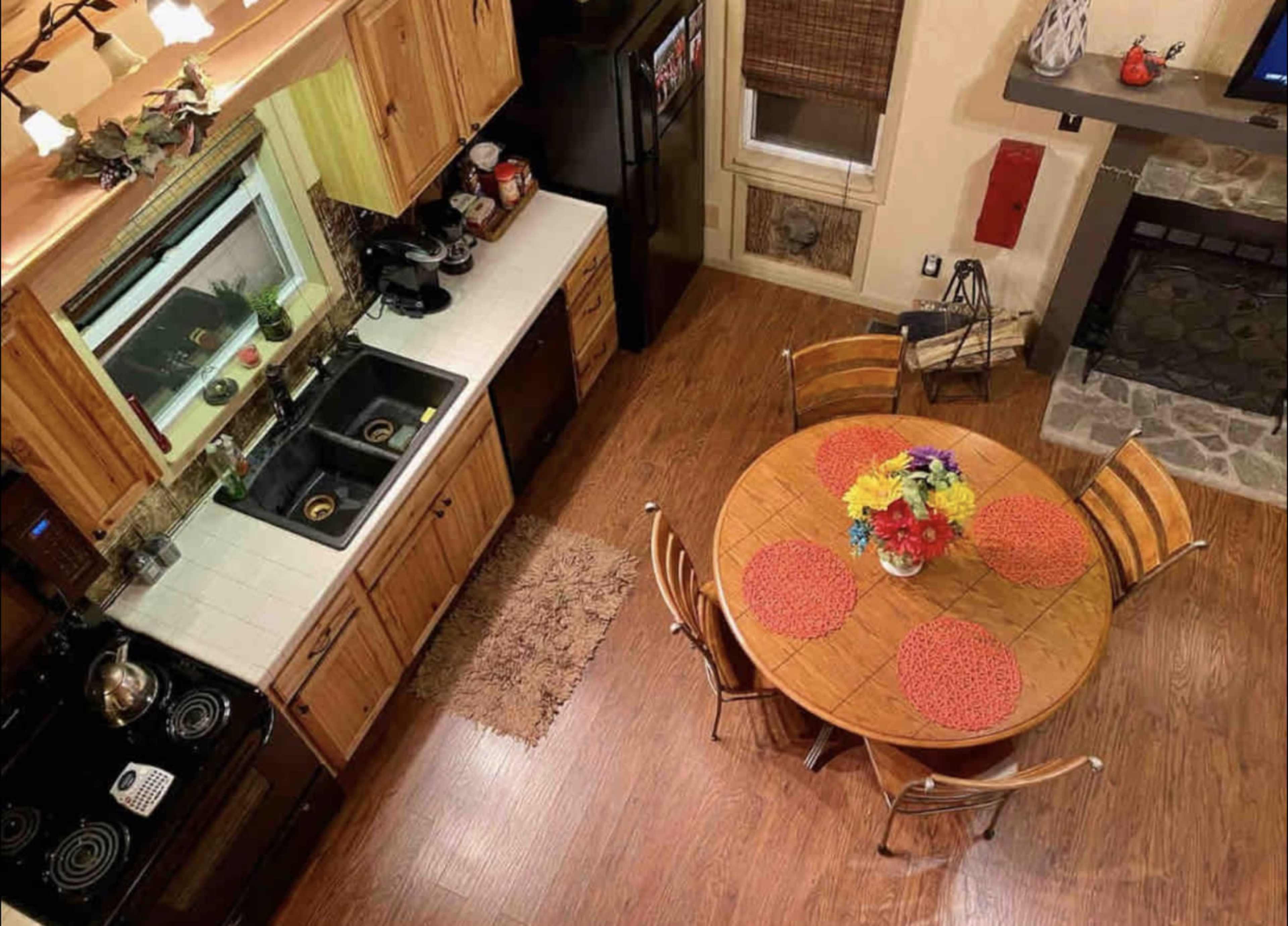 A well-organized kitchen with wooden cabinets, a round dining table set with colorful placemats and flowers, and a cozy seating area near a stone fireplace.