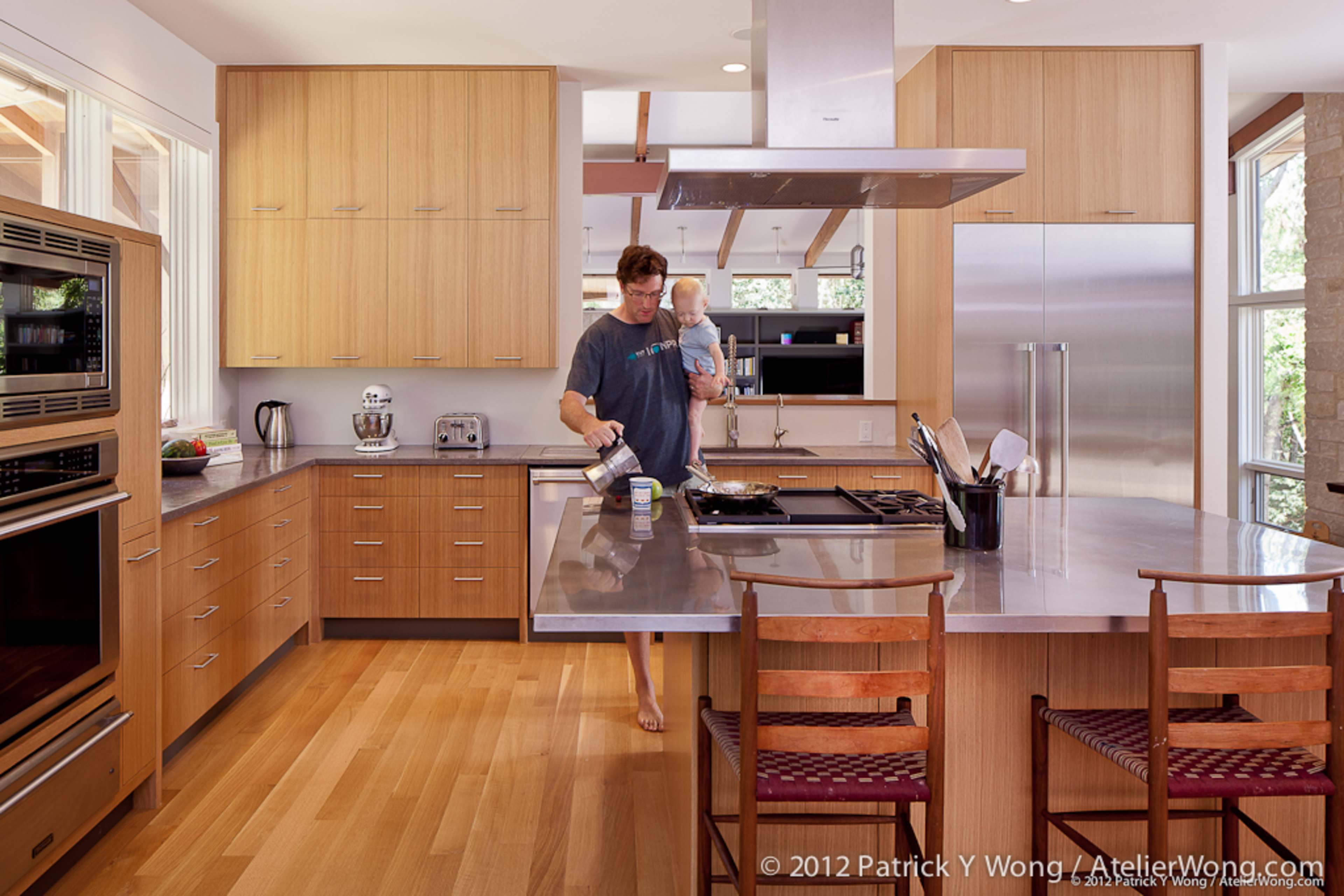 A man stands in a modern kitchen, cooking while holding a small child, with wooden cabinetry and a stainless steel island.