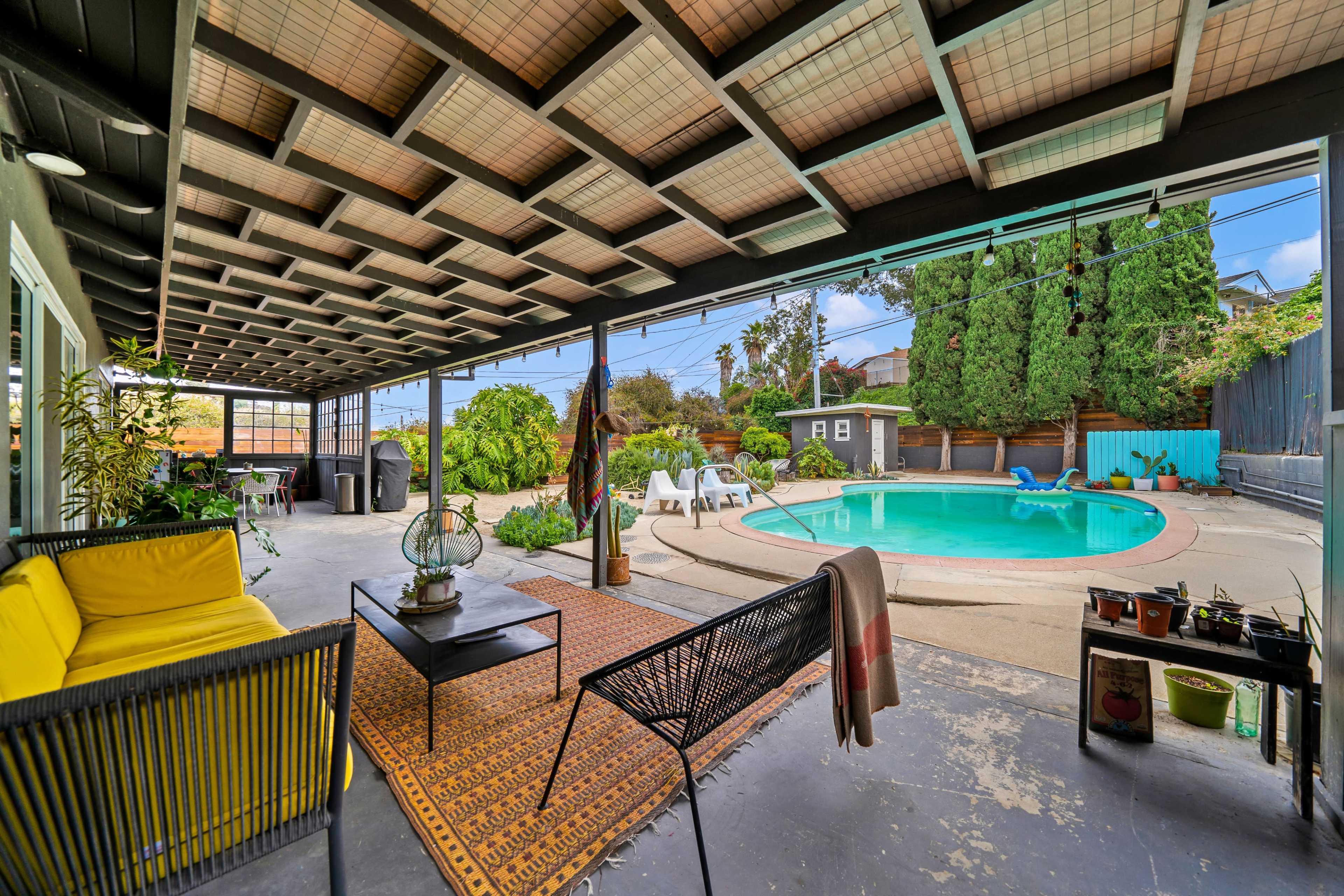 The image shows a covered outdoor patio with seating, a swimming pool in the background, and various plants and decorations surrounding the area.