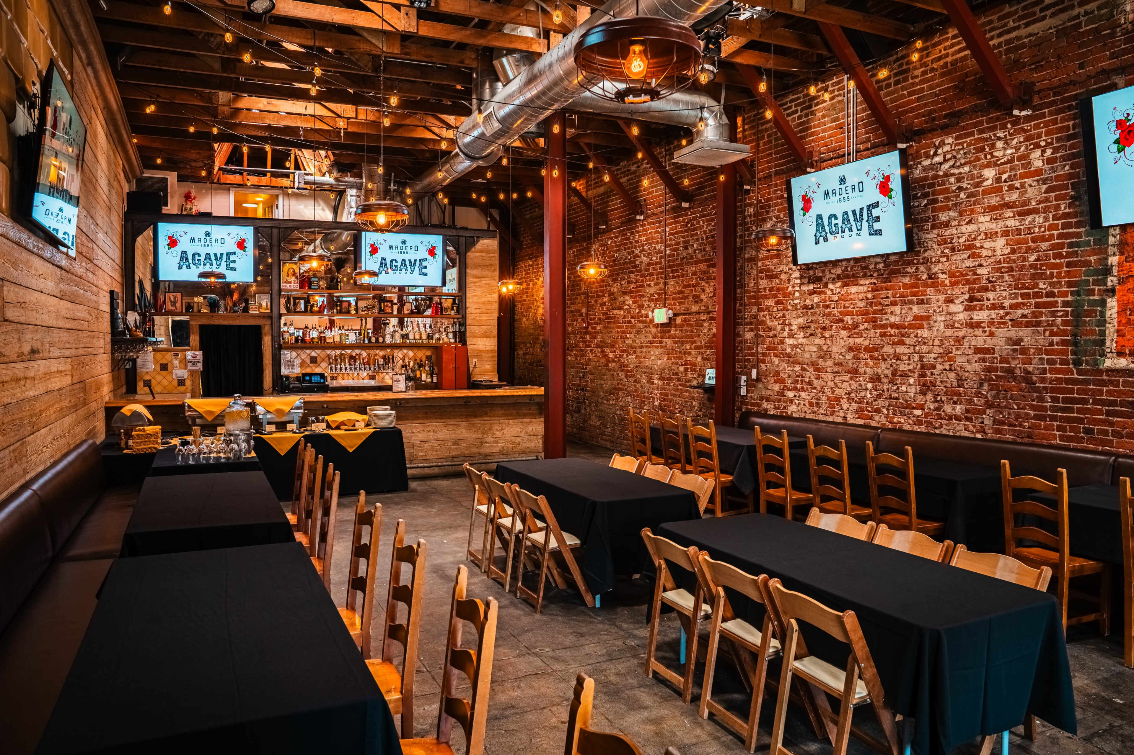 The image shows a rustic restaurant interior featuring wooden tables and chairs arranged facing a bar with multiple screens displaying graphics, illuminated by string lights overhead.