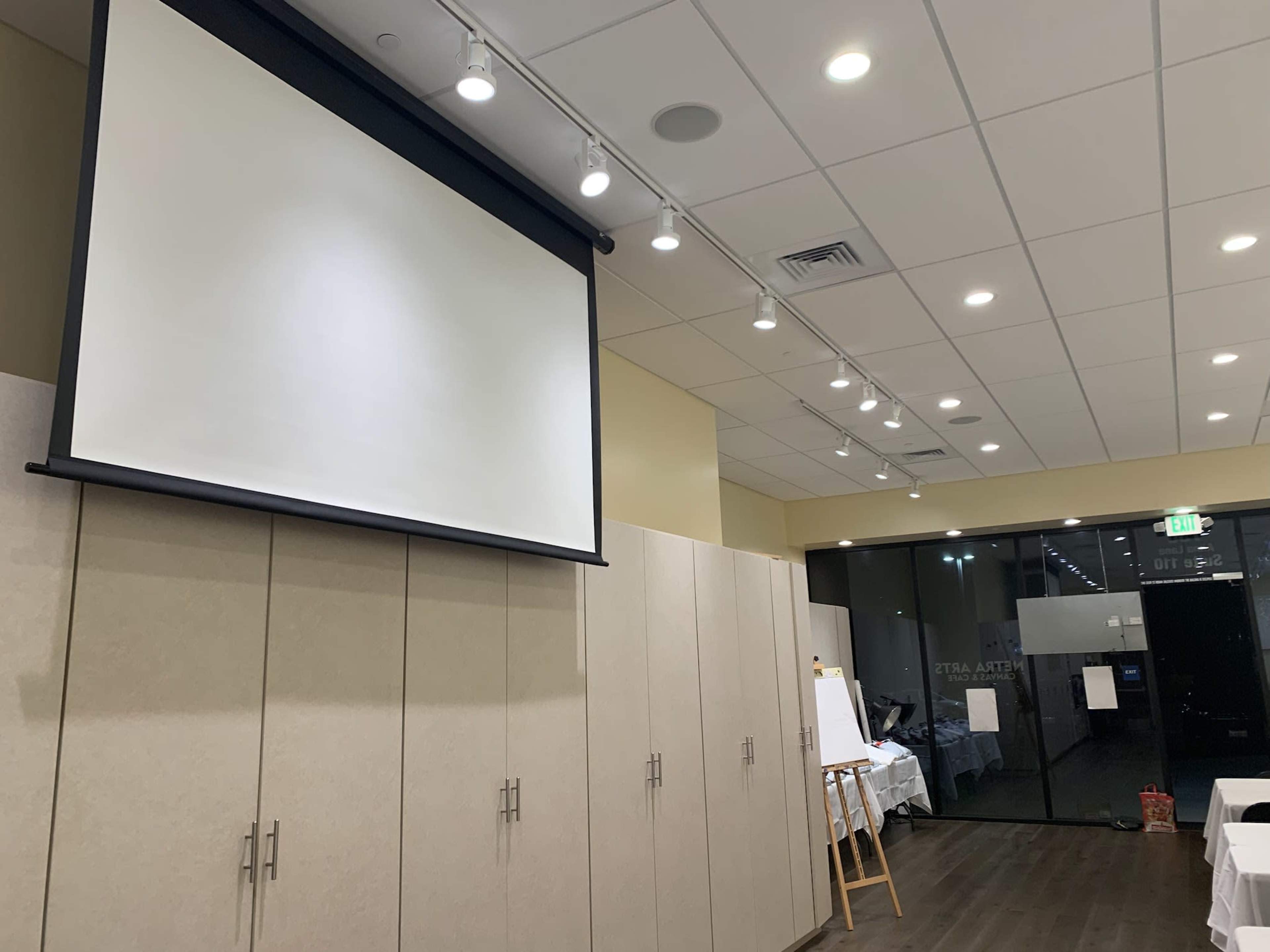 The image depicts a conference room featuring a large projection screen mounted on a wall, with beige cabinetry and bright overhead lighting.