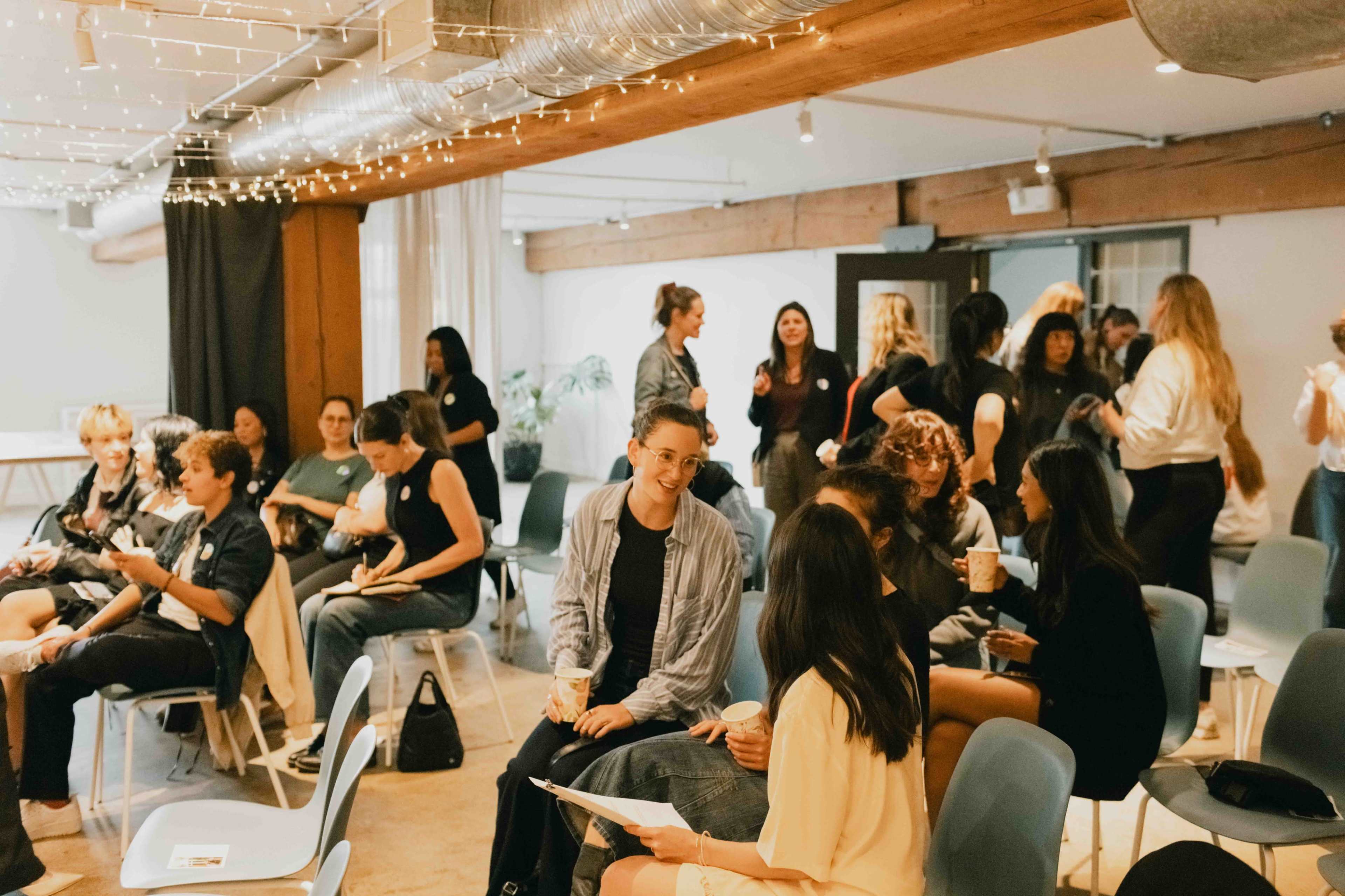 A group of people engages in conversation while seated in a well-lit room with modern decor and string lights.