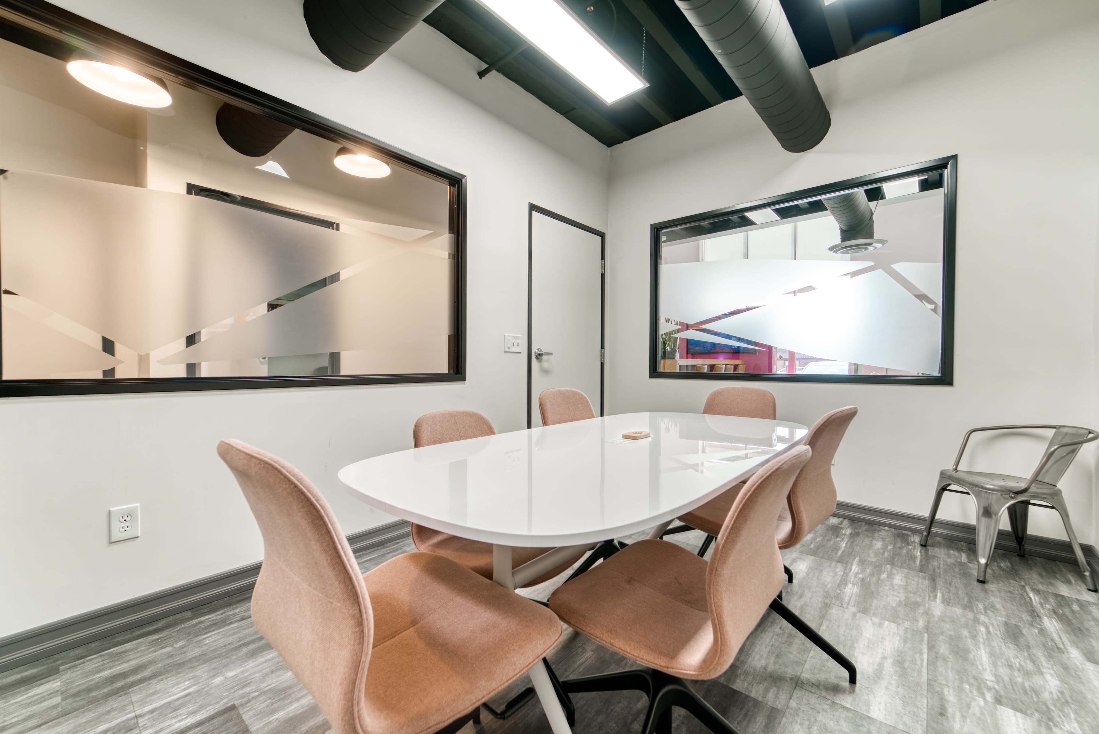 A meeting room features a white oval table surrounded by four upholstered chairs, with two large windows displaying frosted glass.