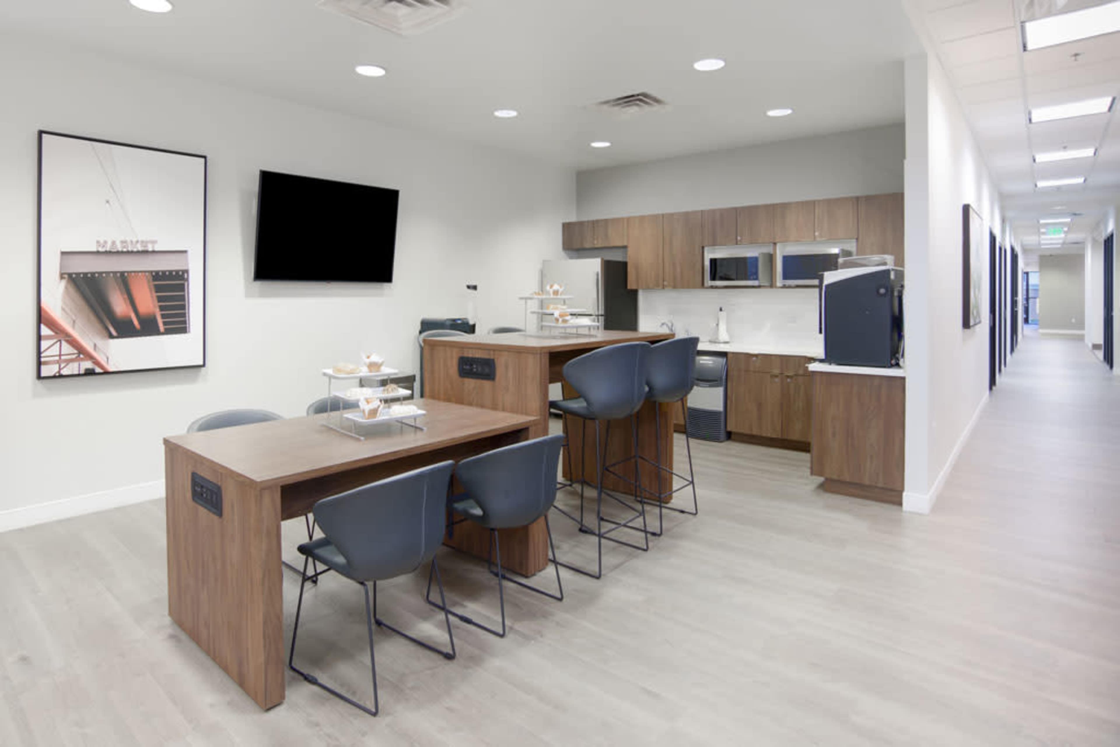 A modern kitchenette with wooden tables and gray chairs, accompanied by a refrigerator, microwave, and sink, along a well-lit corridor.