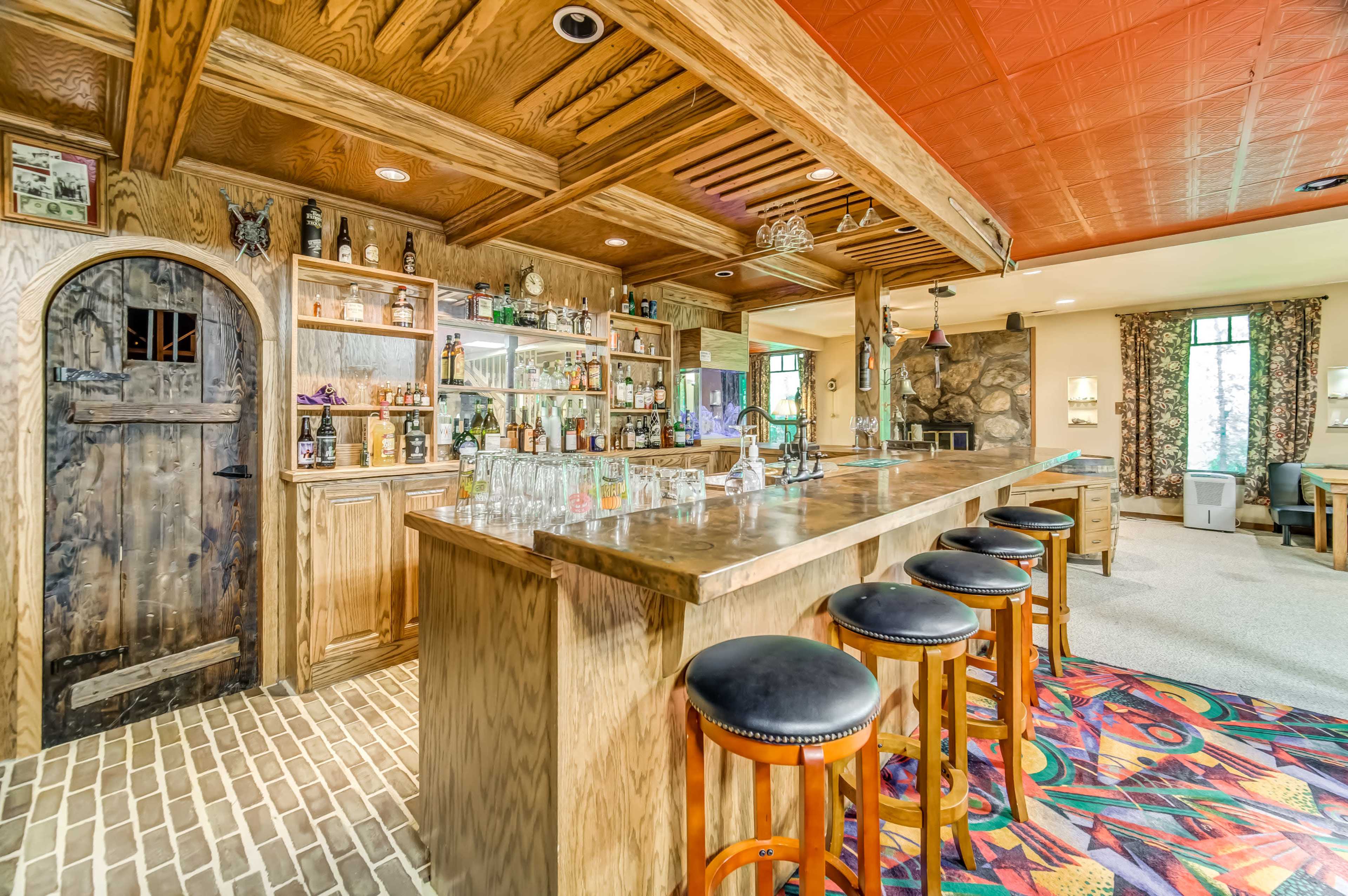 A wooden bar area features a large counter with several stools, shelves stocked with bottles, and a stone wall backdrop in a well-lit room.