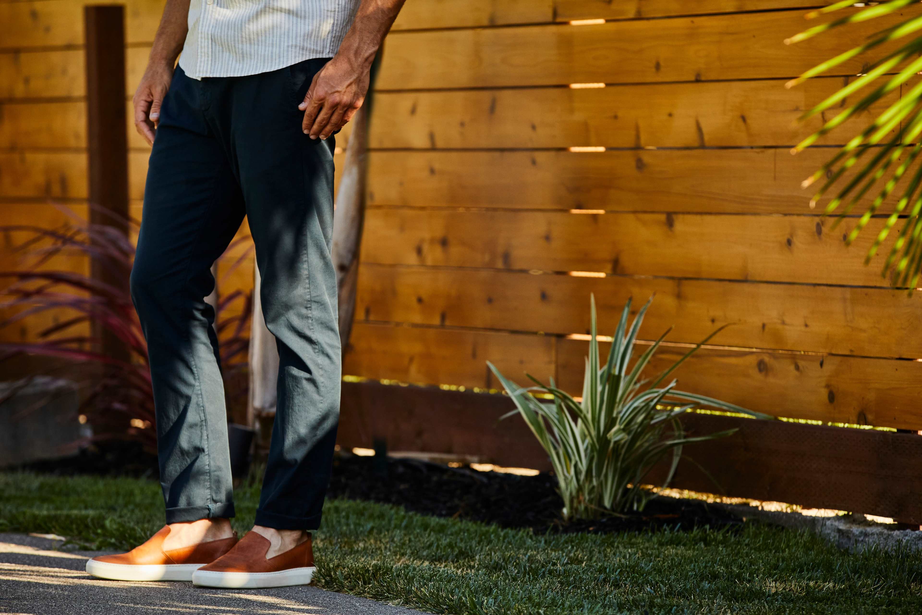 A man stands outdoors next to a wooden fence, wearing a light shirt and brown slip-on shoes, with grass and plants visible nearby.