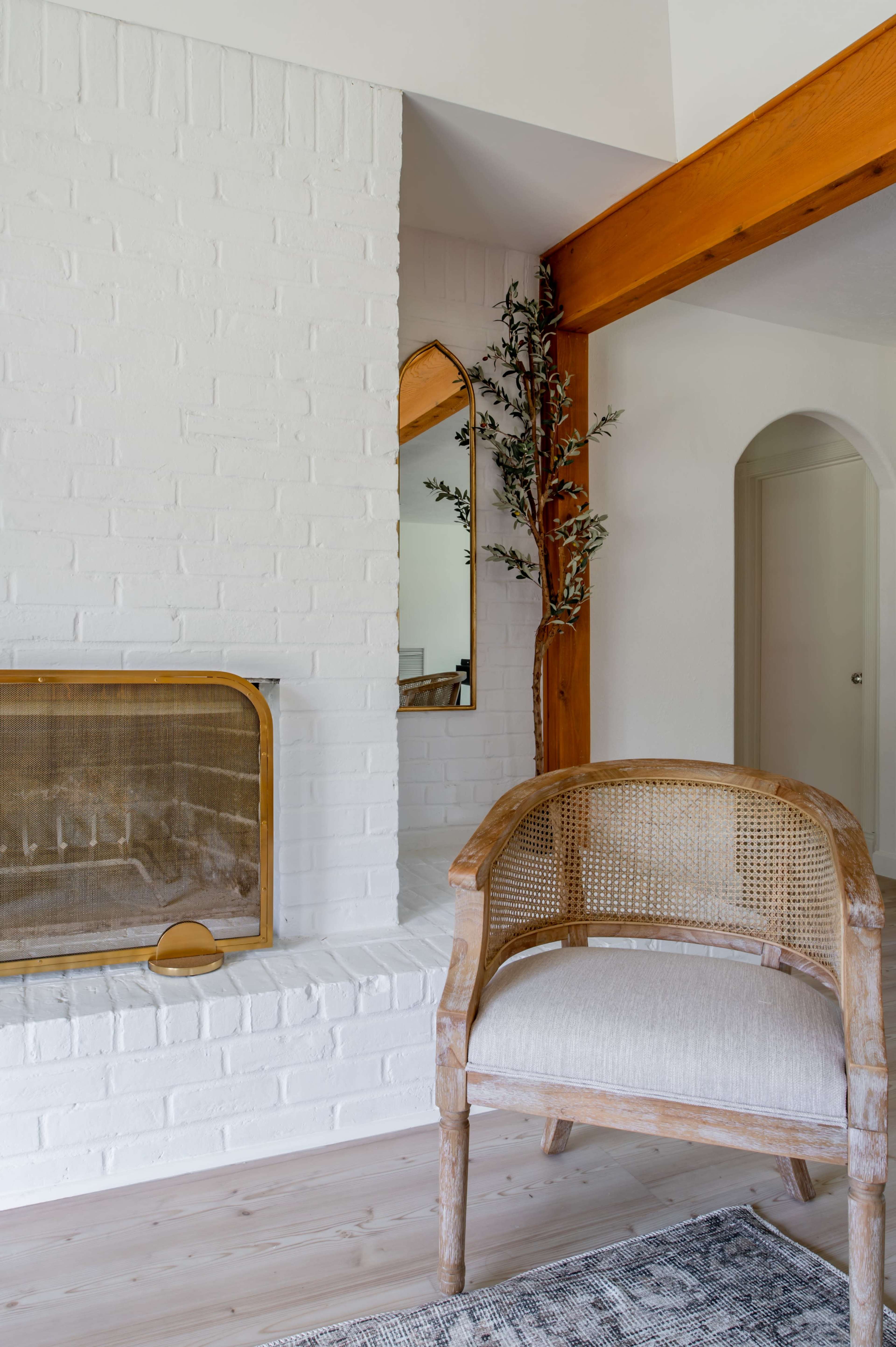 A light-filled living room featuring a brick fireplace, a wooden arched mirror, a decorative plant, and a cane-backed chair.