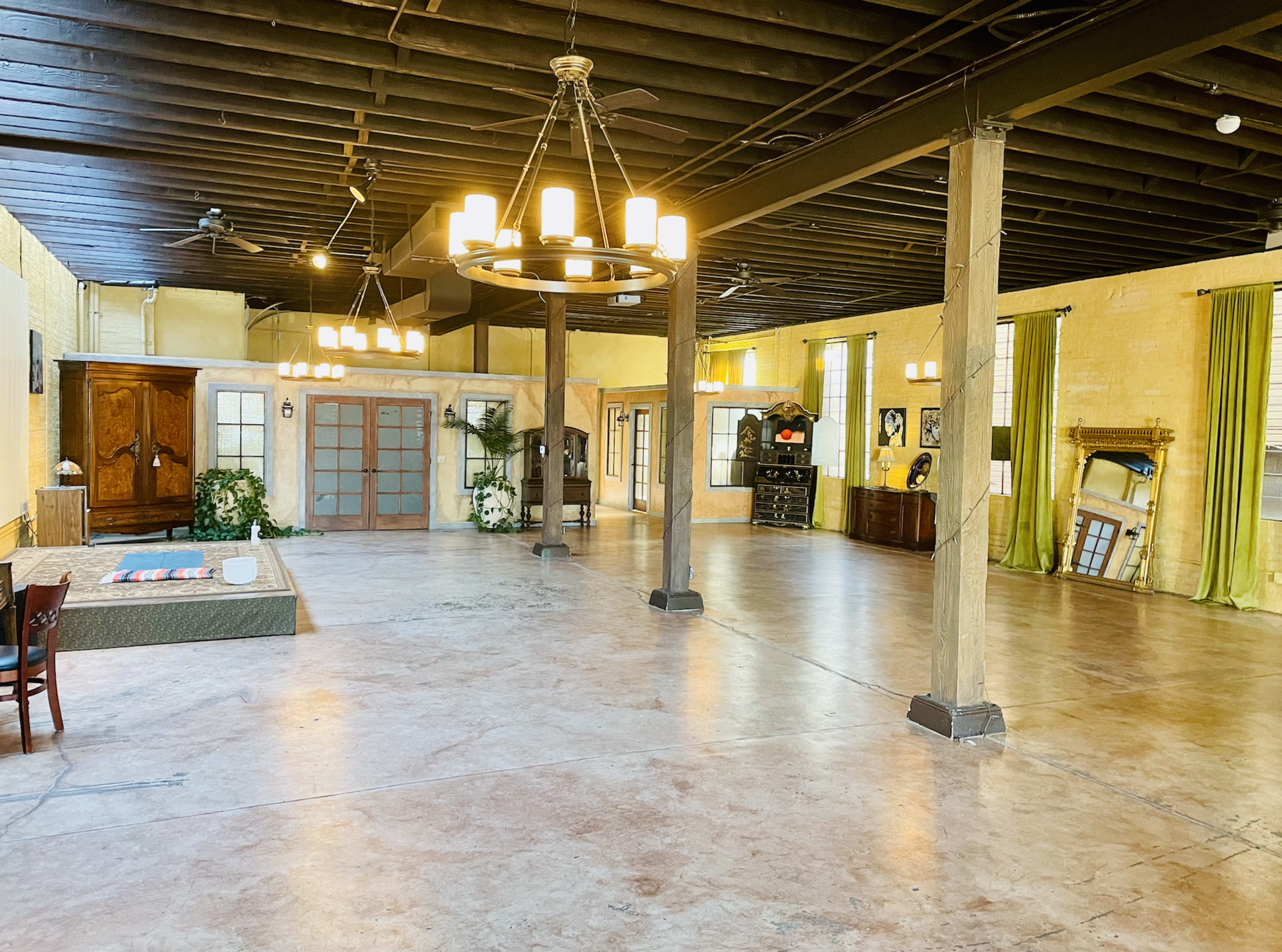 A large, open room with polished concrete floors, exposed beams, and chandeliers, featuring wooden furniture and decorative elements along the walls.