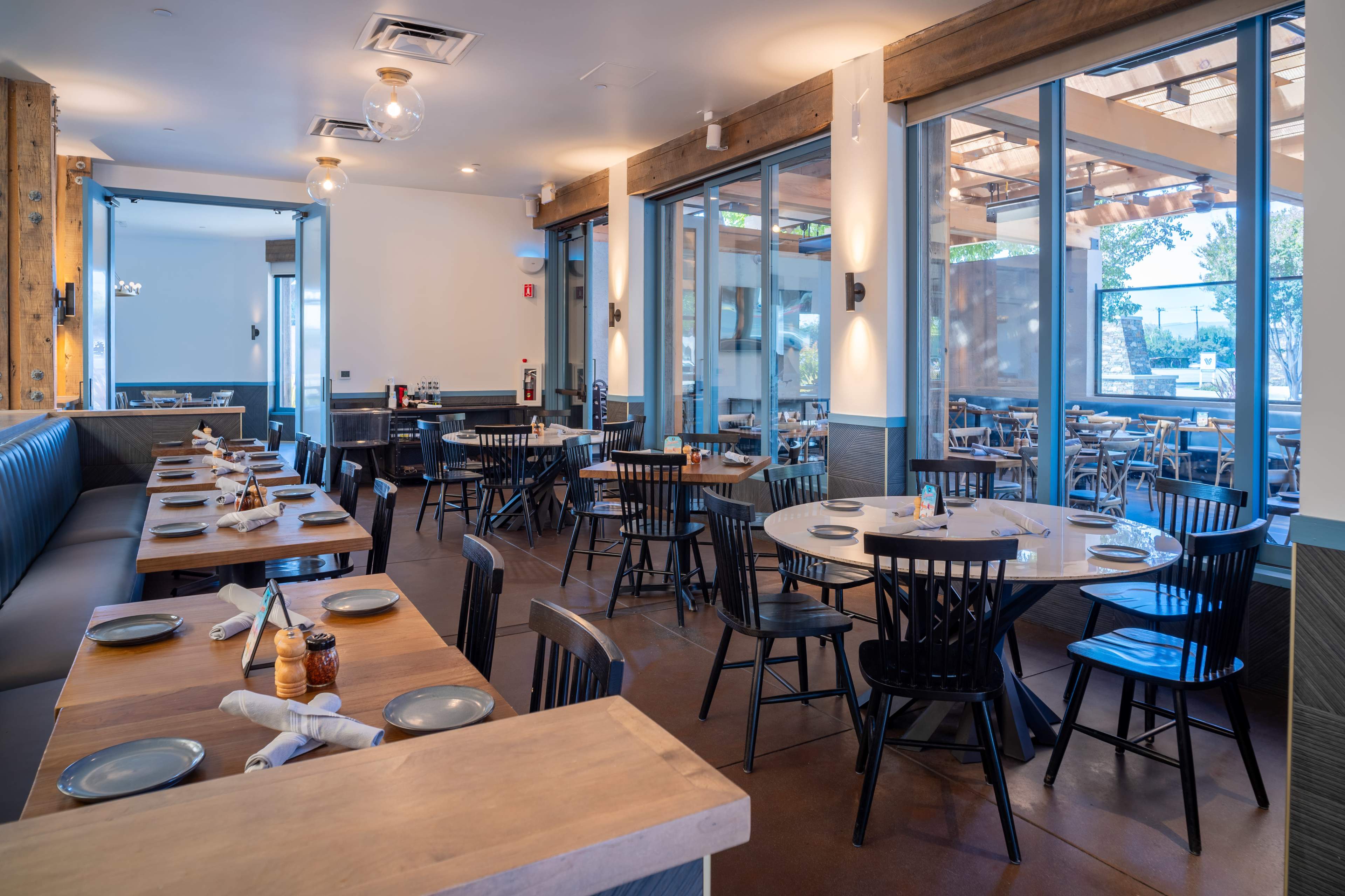 The image shows a modern restaurant interior with multiple tables and chairs arranged in a spacious layout, featuring large windows and wooden accents.