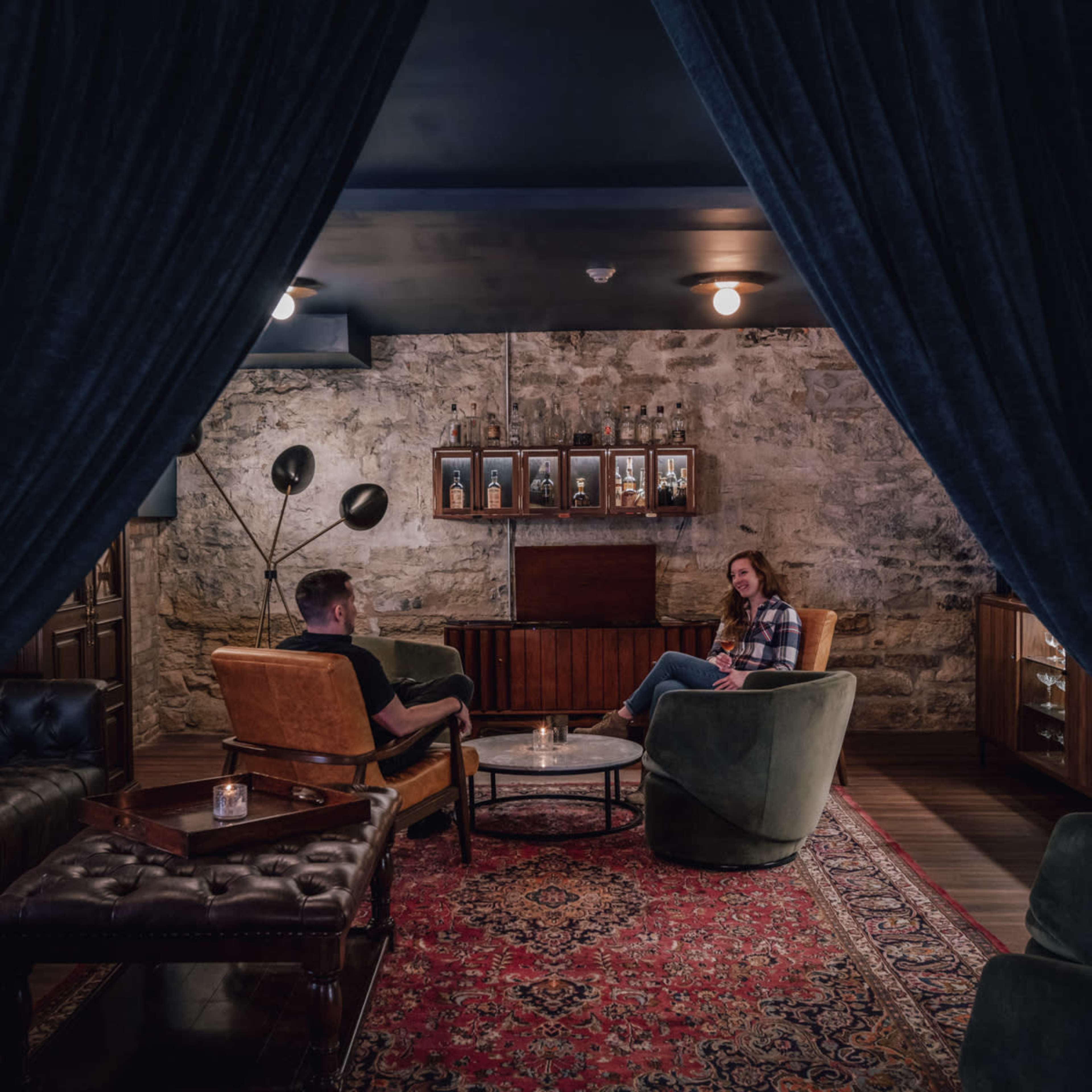 Two people sit in armchairs across from each other in a dimly lit lounge with exposed stone walls and a patterned rug.