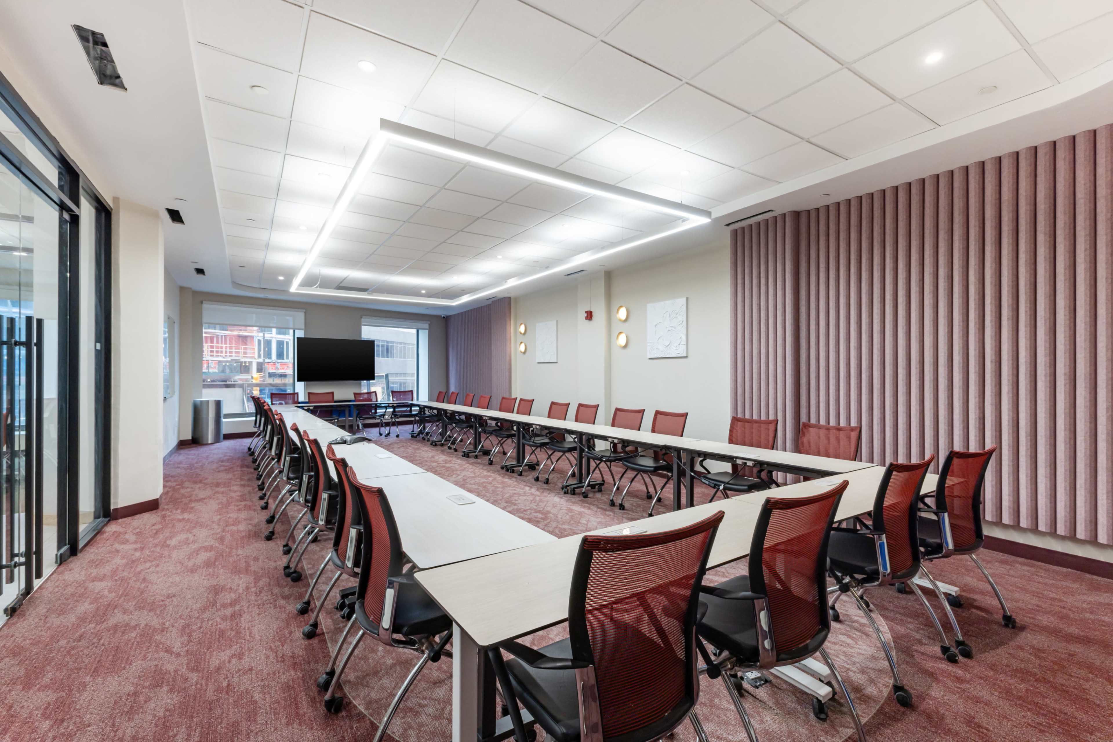 The image shows a modern conference room with a long table arranged in a U-shape, surrounded by ergonomic chairs, and equipped with a large screen at one end.