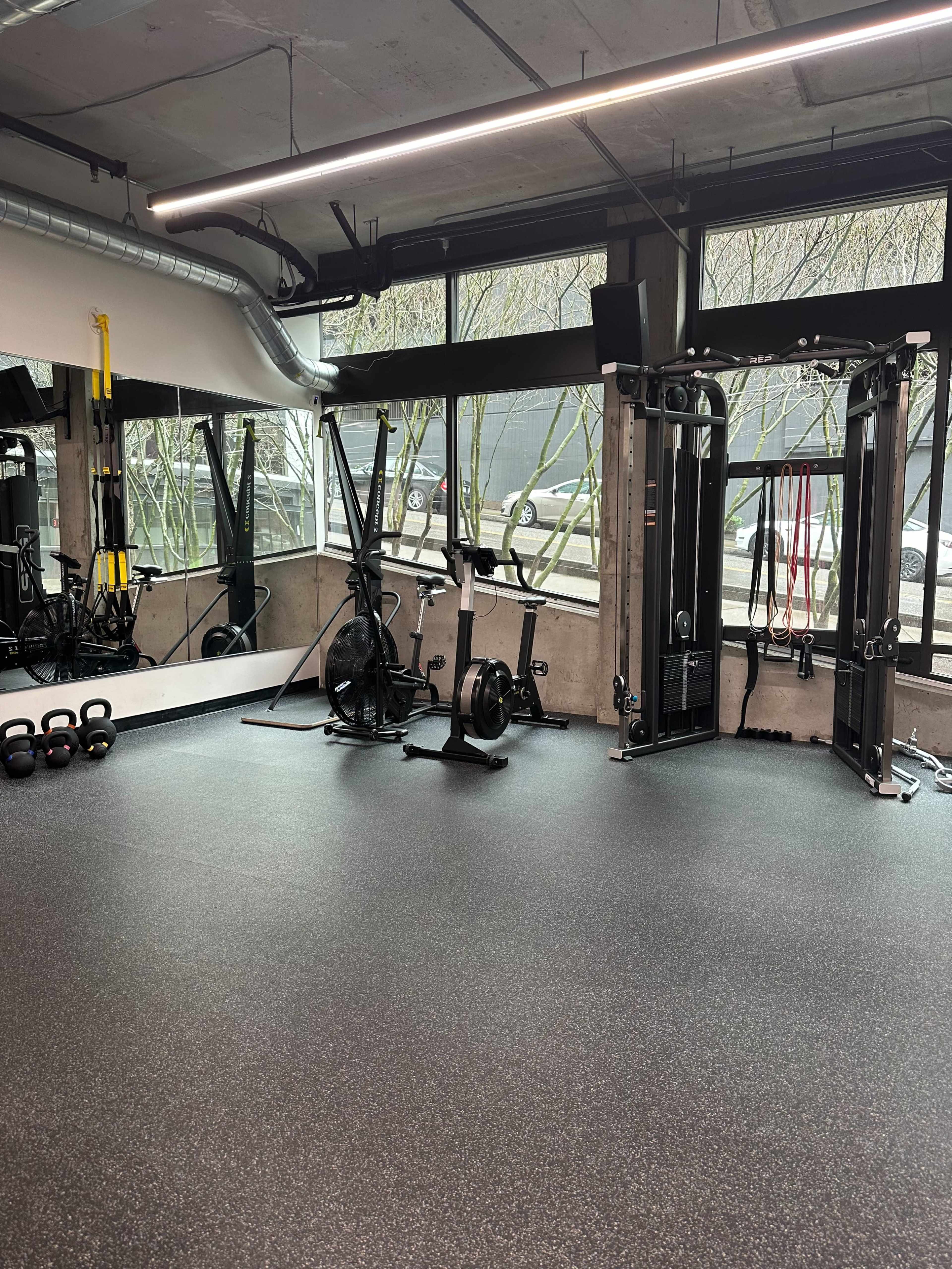 A fitness studio equipped with various exercise machines, weights, and large windows allowing natural light to enter.