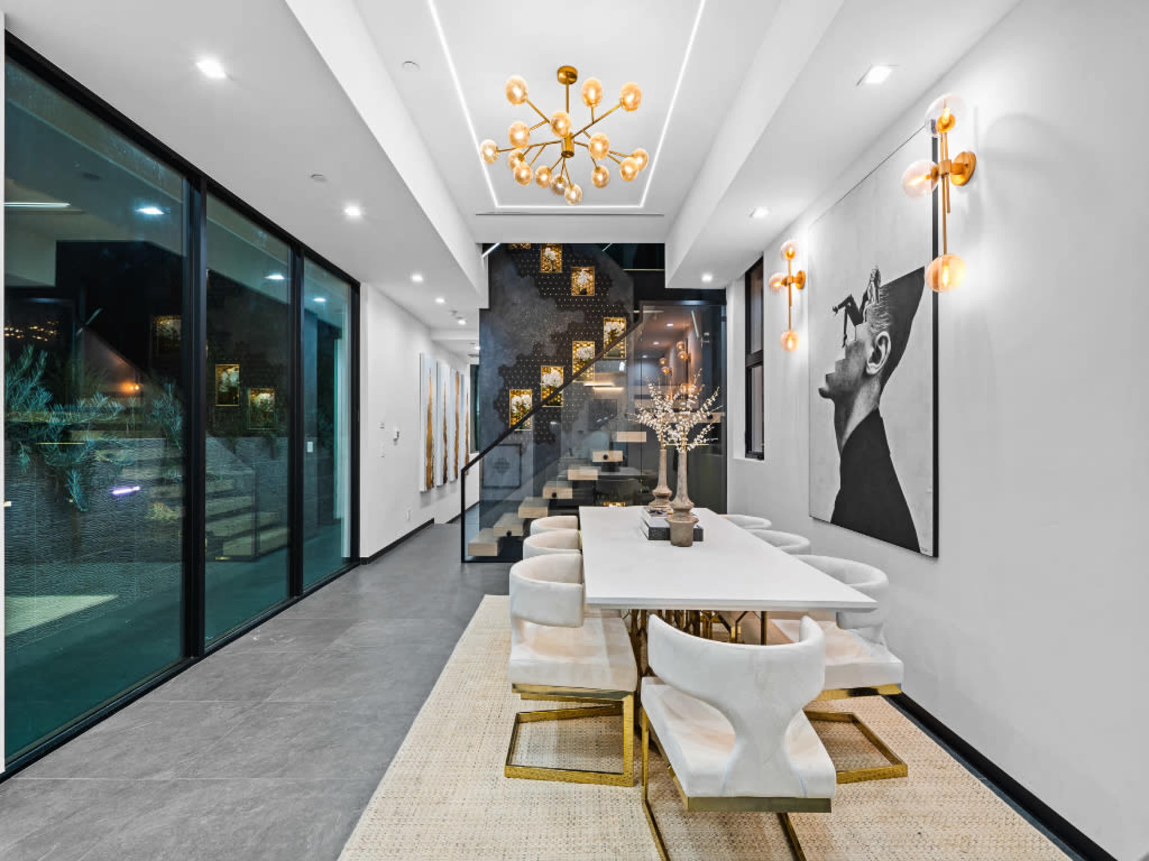 The image features a modern dining area with a large table surrounded by white chairs, adjacent to a staircase and large windows.