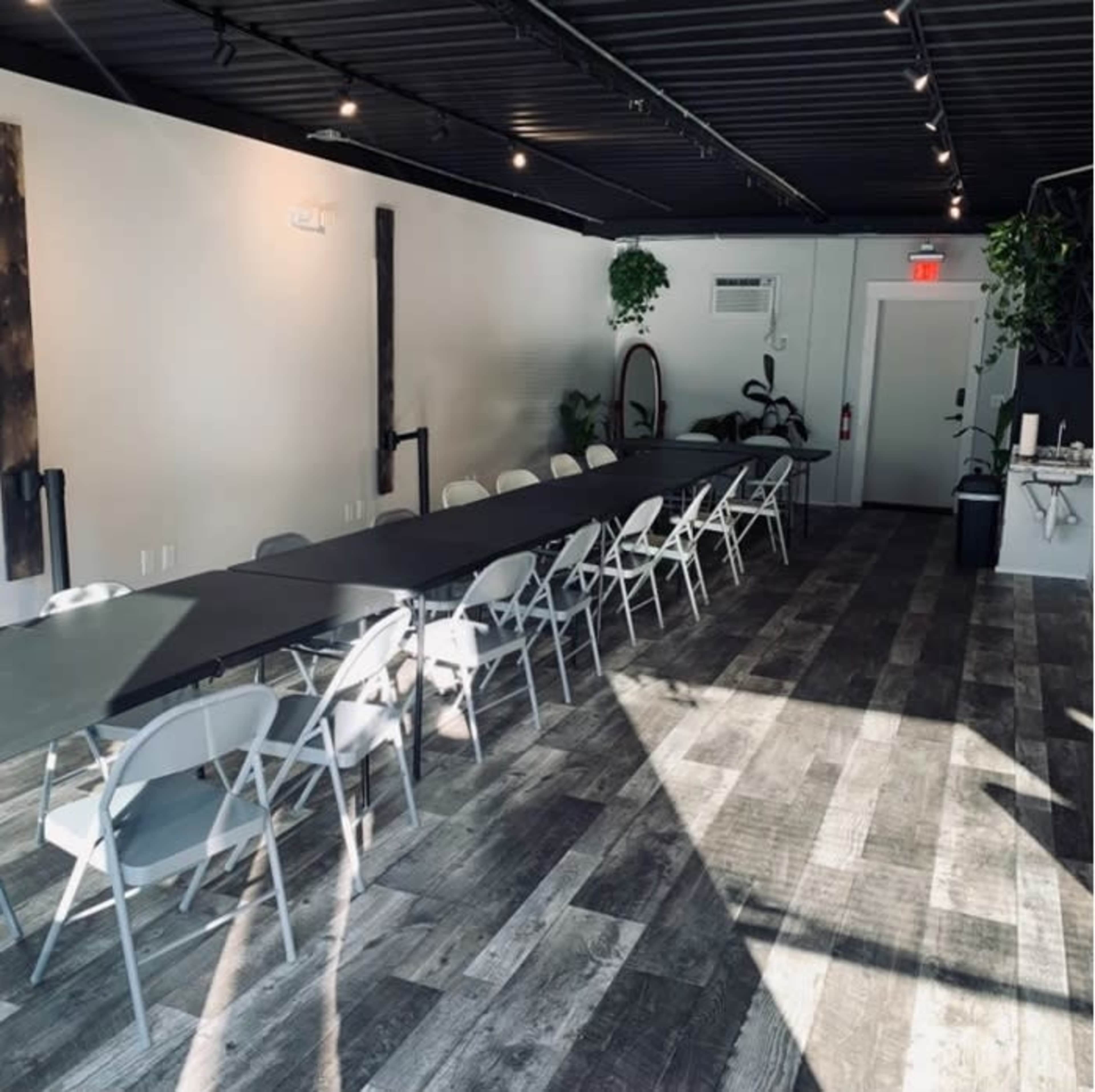 A long, rectangular table with white chairs is set up in a well-lit, modern meeting room featuring wood flooring and green plants.