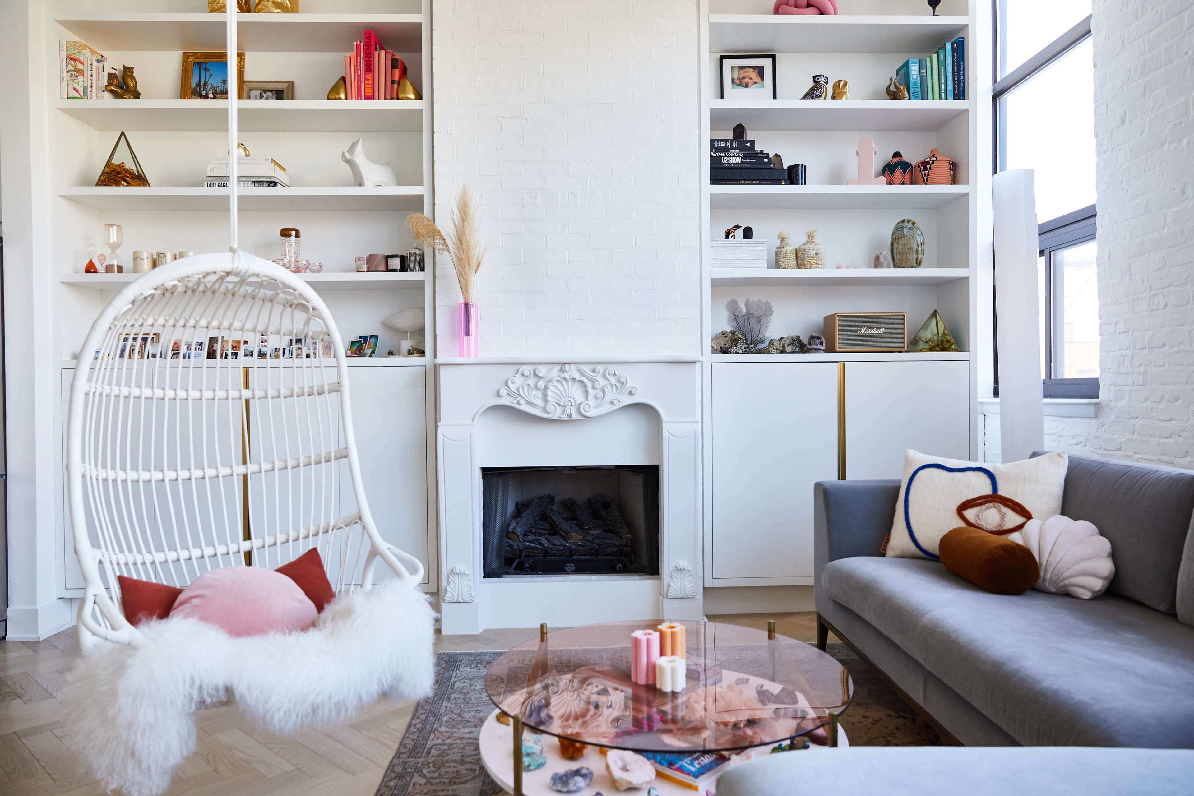 The image shows a modern living room with a white hanging chair, a decorative fireplace, and shelves filled with various objects and books.