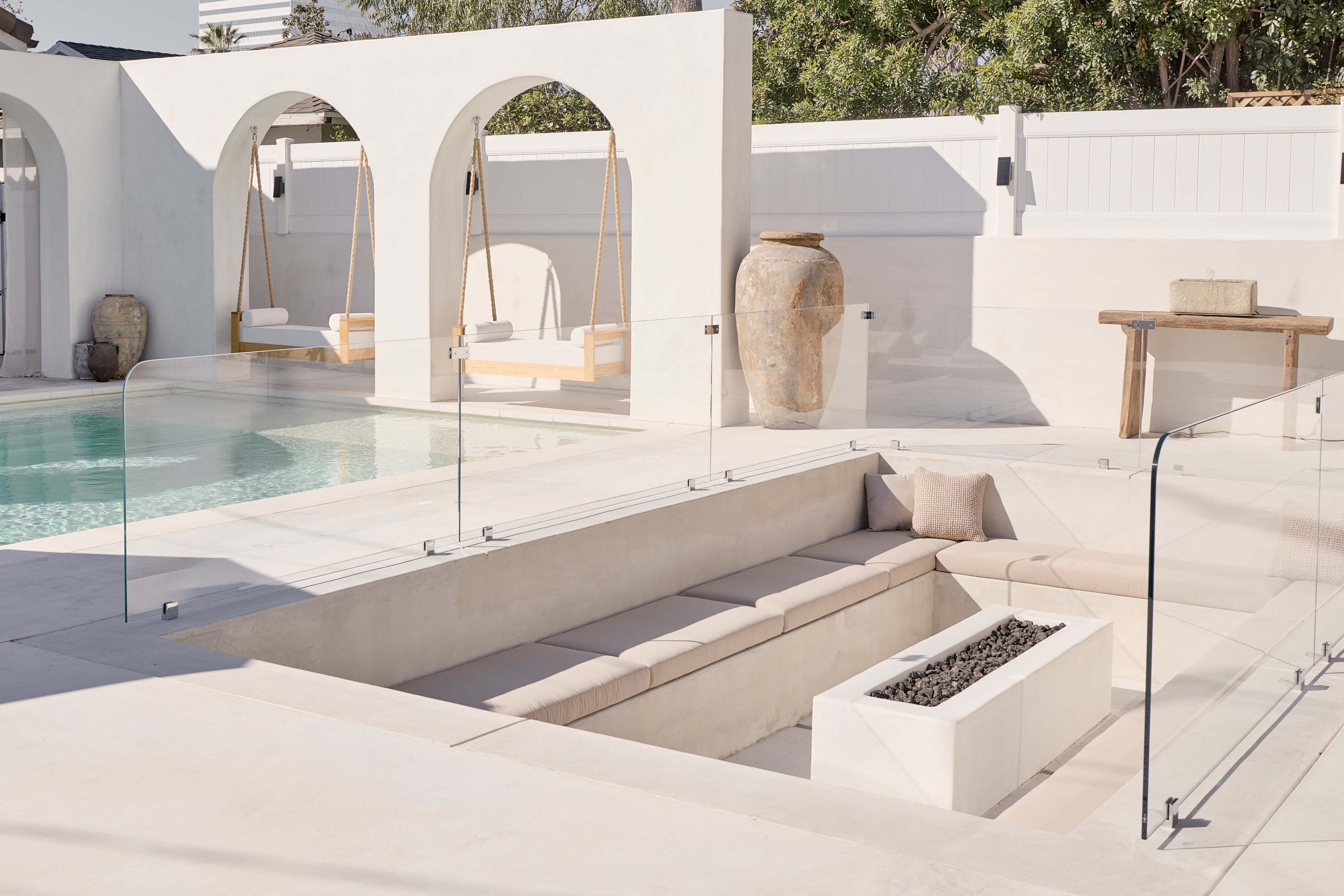 The image features a modern outdoor space with a swimming pool, two swing chairs under arched structures, a stone table, and a seating area with a sunken fire pit surrounded by clear glass barriers.