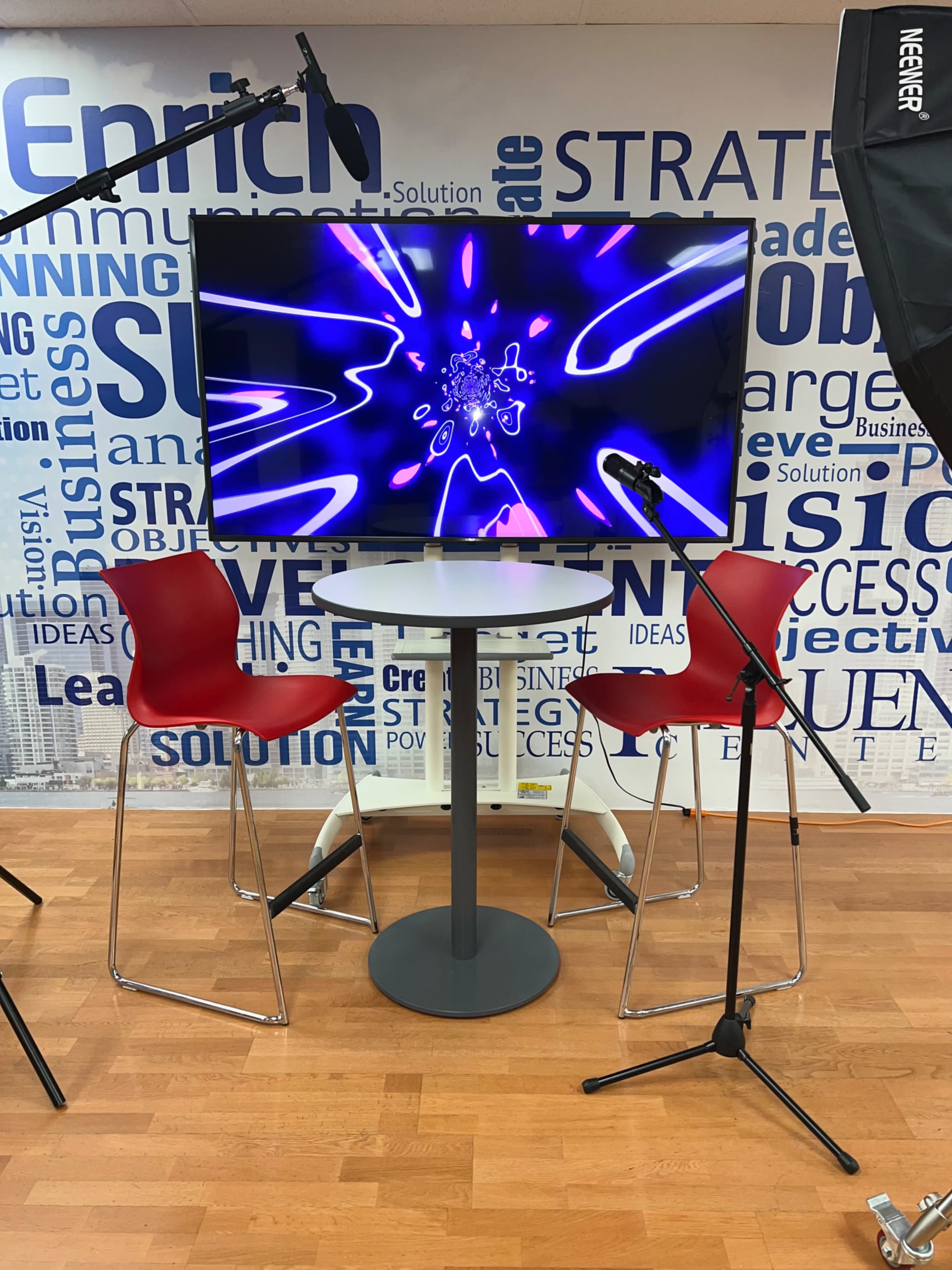 A modern display setup features a large screen with vibrant visuals, surrounded by two red chairs and a circular table, positioned in a well-lit room decorated with various business-related words.