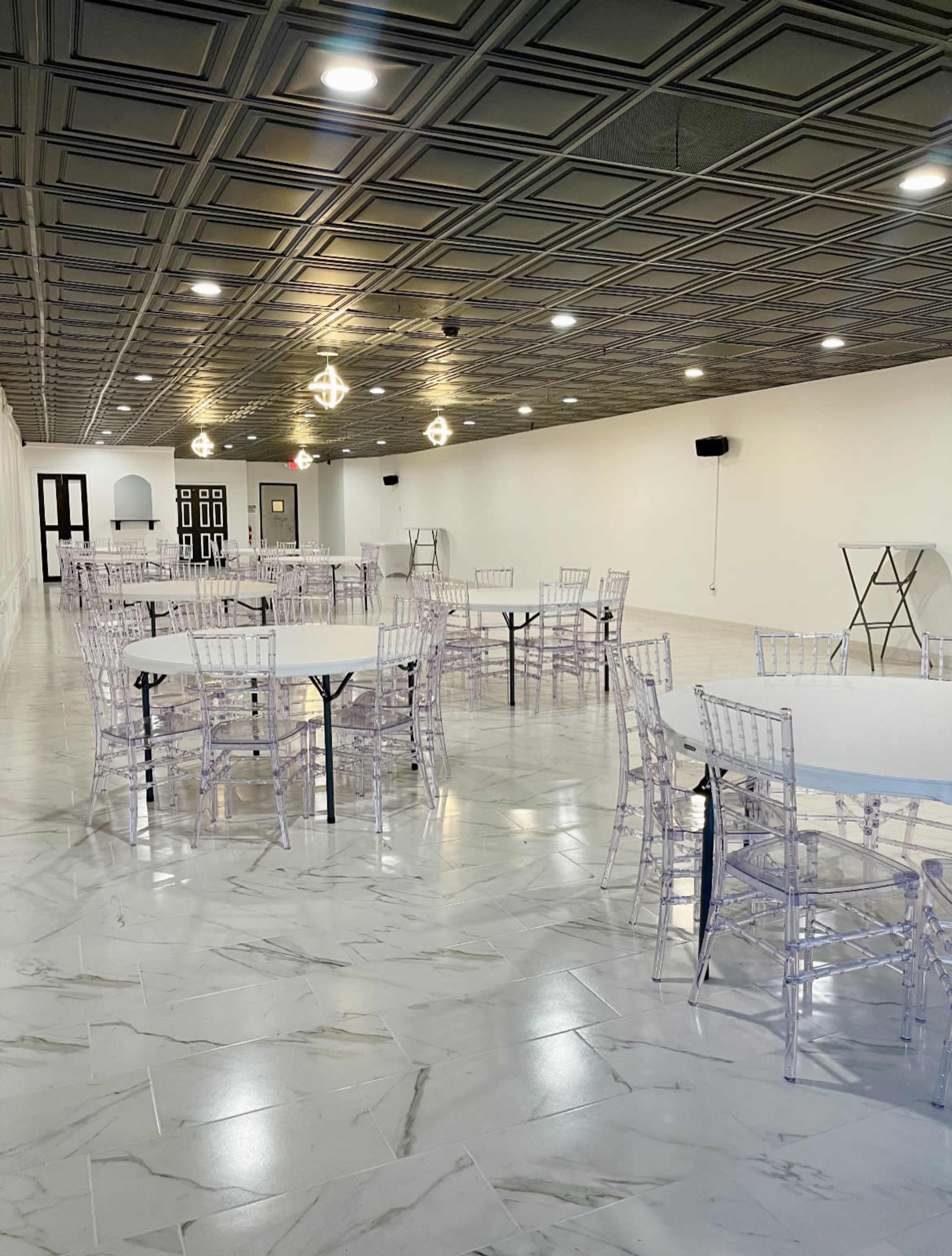 A spacious event room with several transparent chairs arranged around white tables on a marble floor.