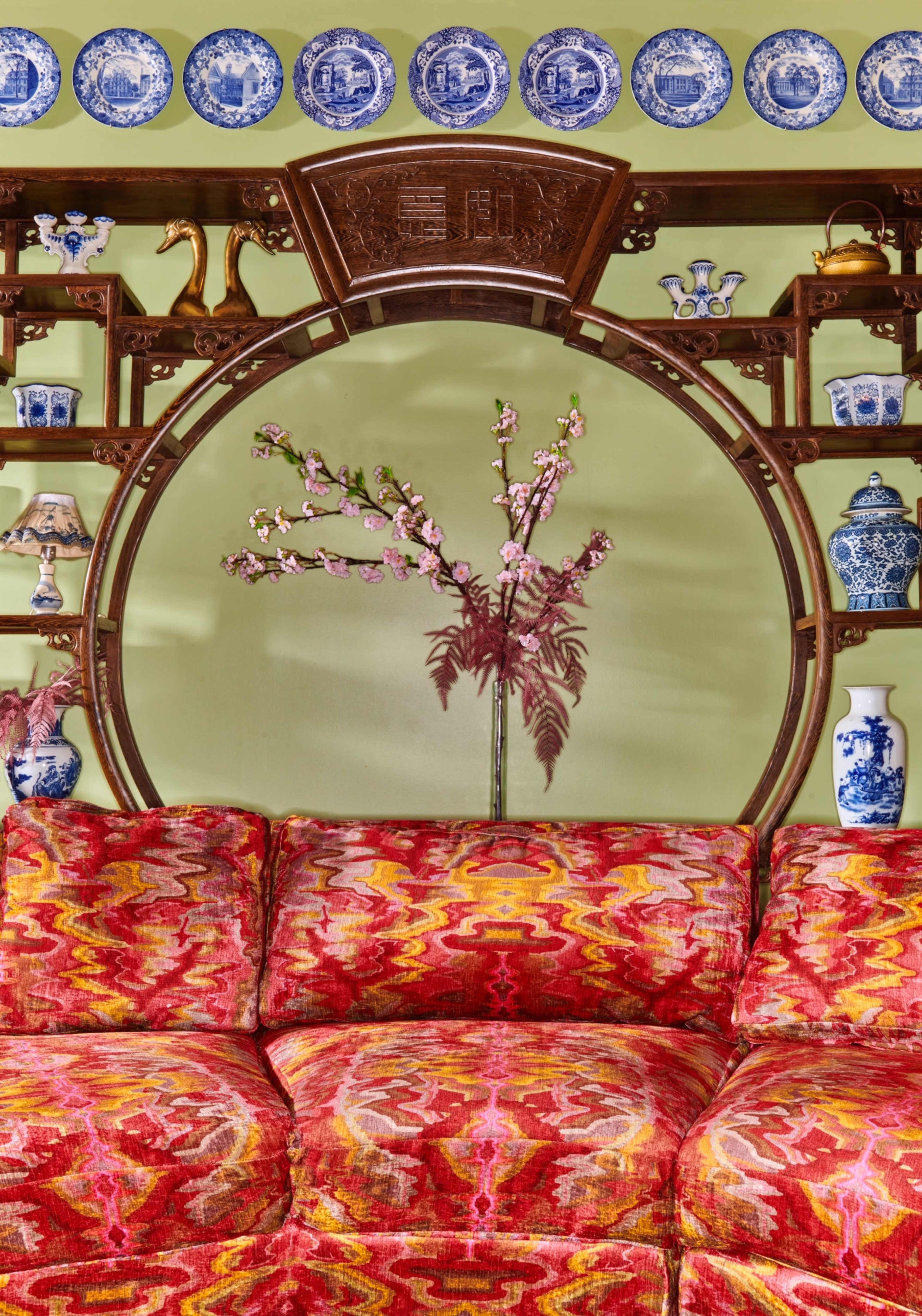 A vibrant red and orange patterned sofa in front of a circular wooden shelf displaying blue and white porcelain items and a floral arrangement.
