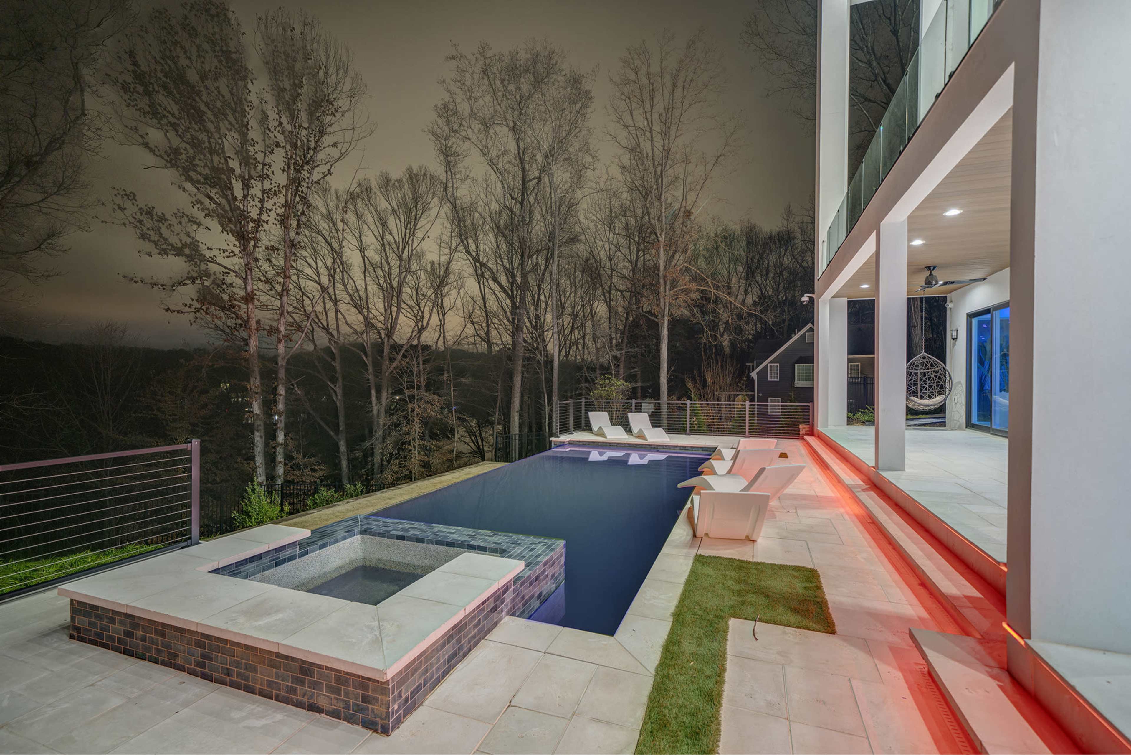 The image shows a modern house with a sleek pool and spa area, surrounded by tall trees under a night sky.
