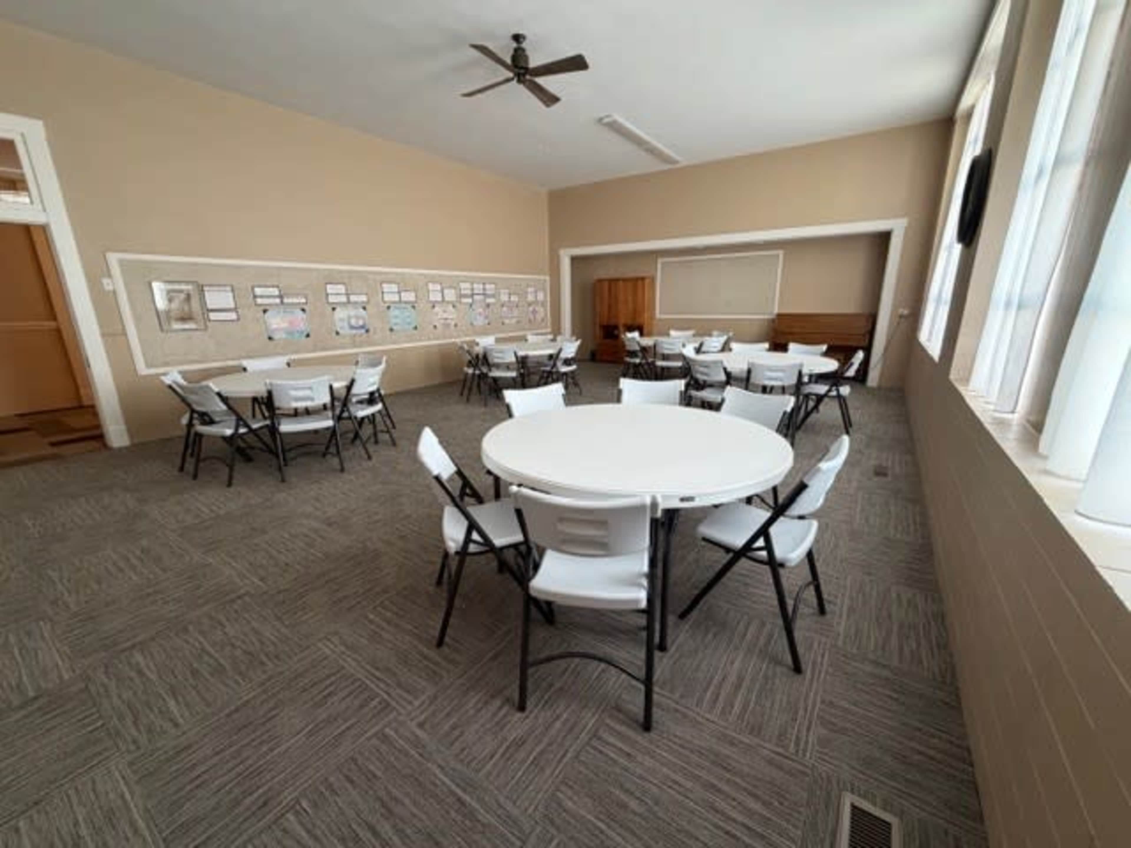 The image shows a spacious room with multiple round tables and chairs arranged, along with a wall displaying various documents or images.