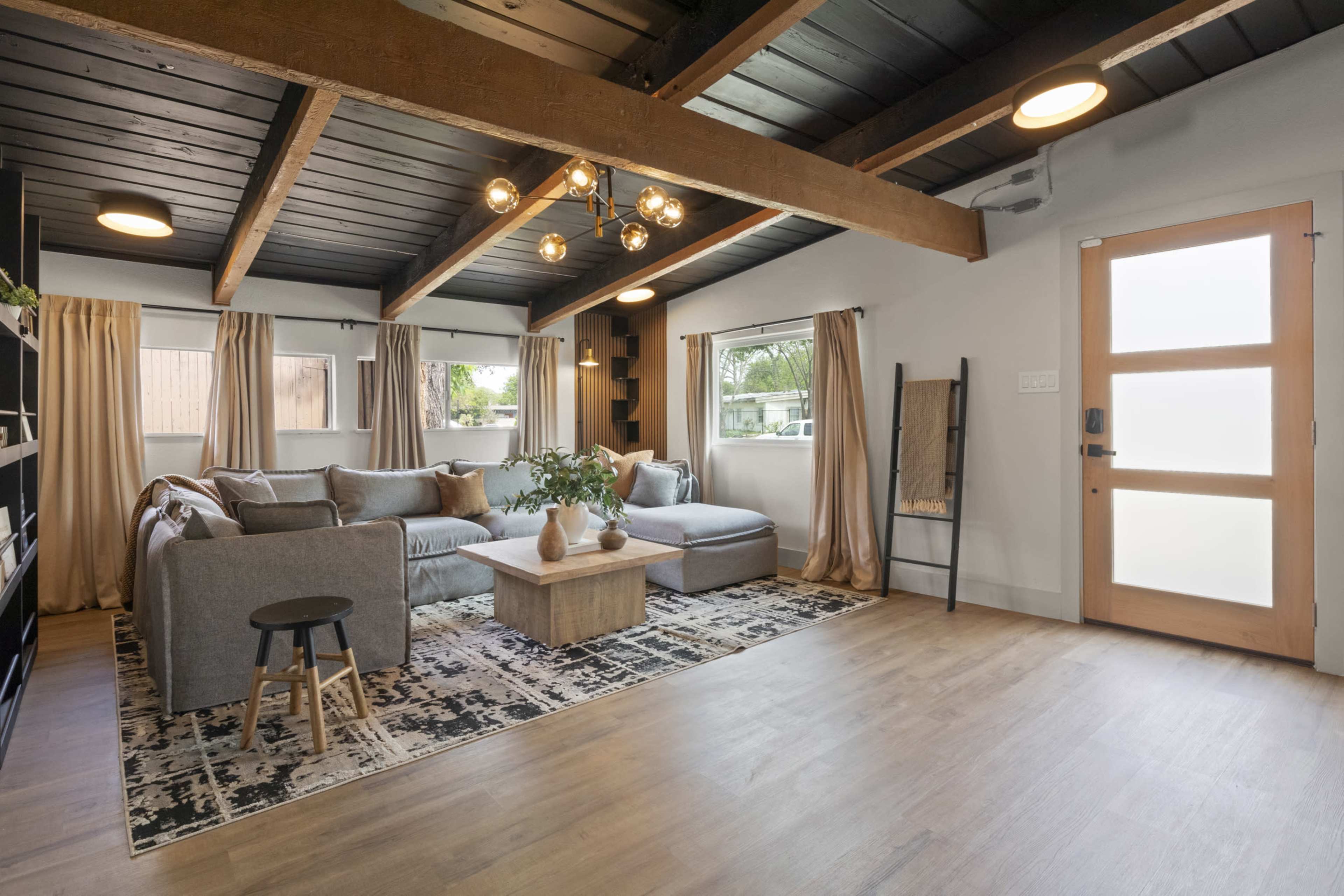 The image features a modern living room with a gray sectional sofa, a light wood coffee table, large windows, and a front door with glass panels.