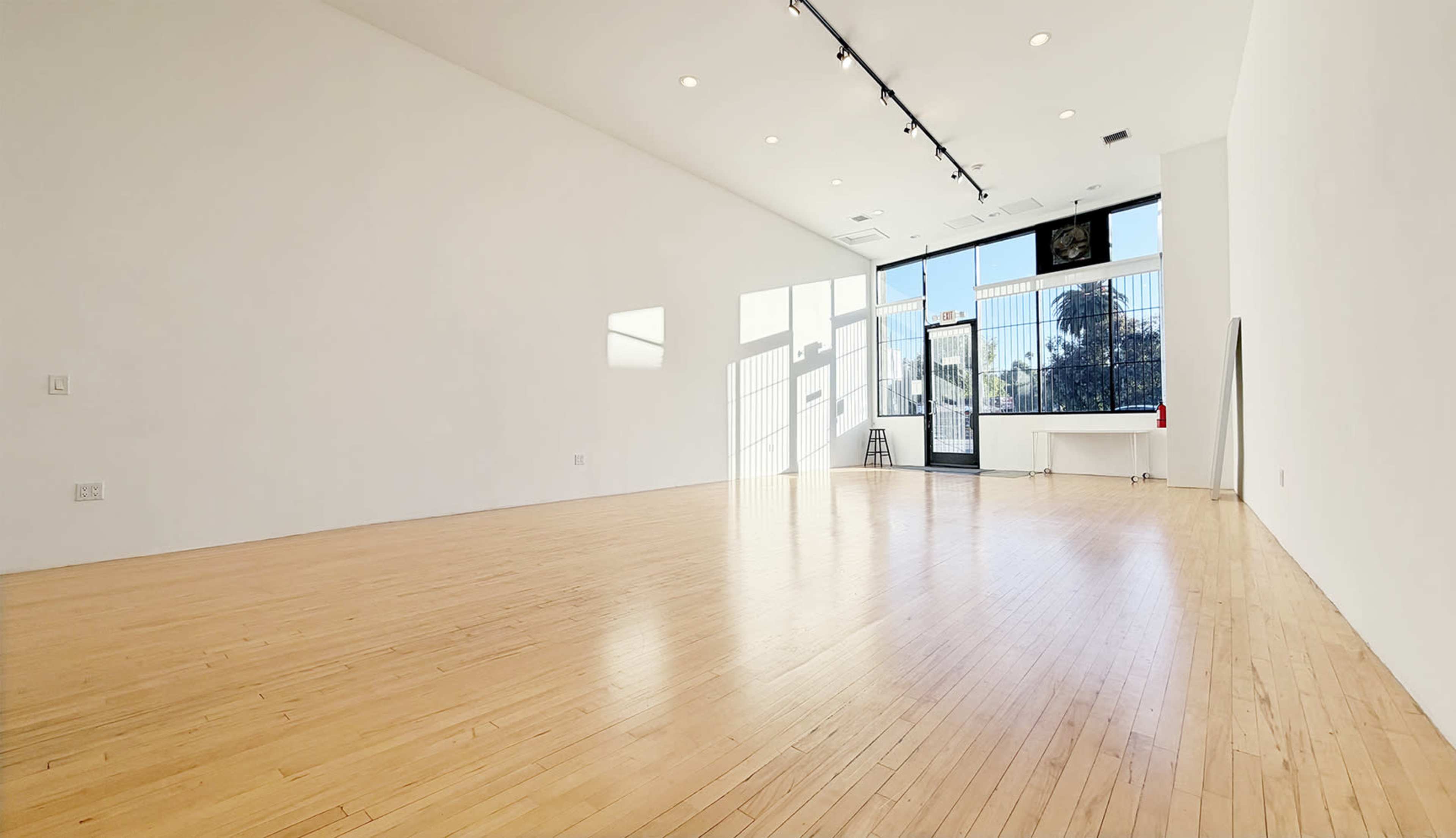 The image shows a spacious, empty room with wooden floors and large windows allowing natural light to enter.