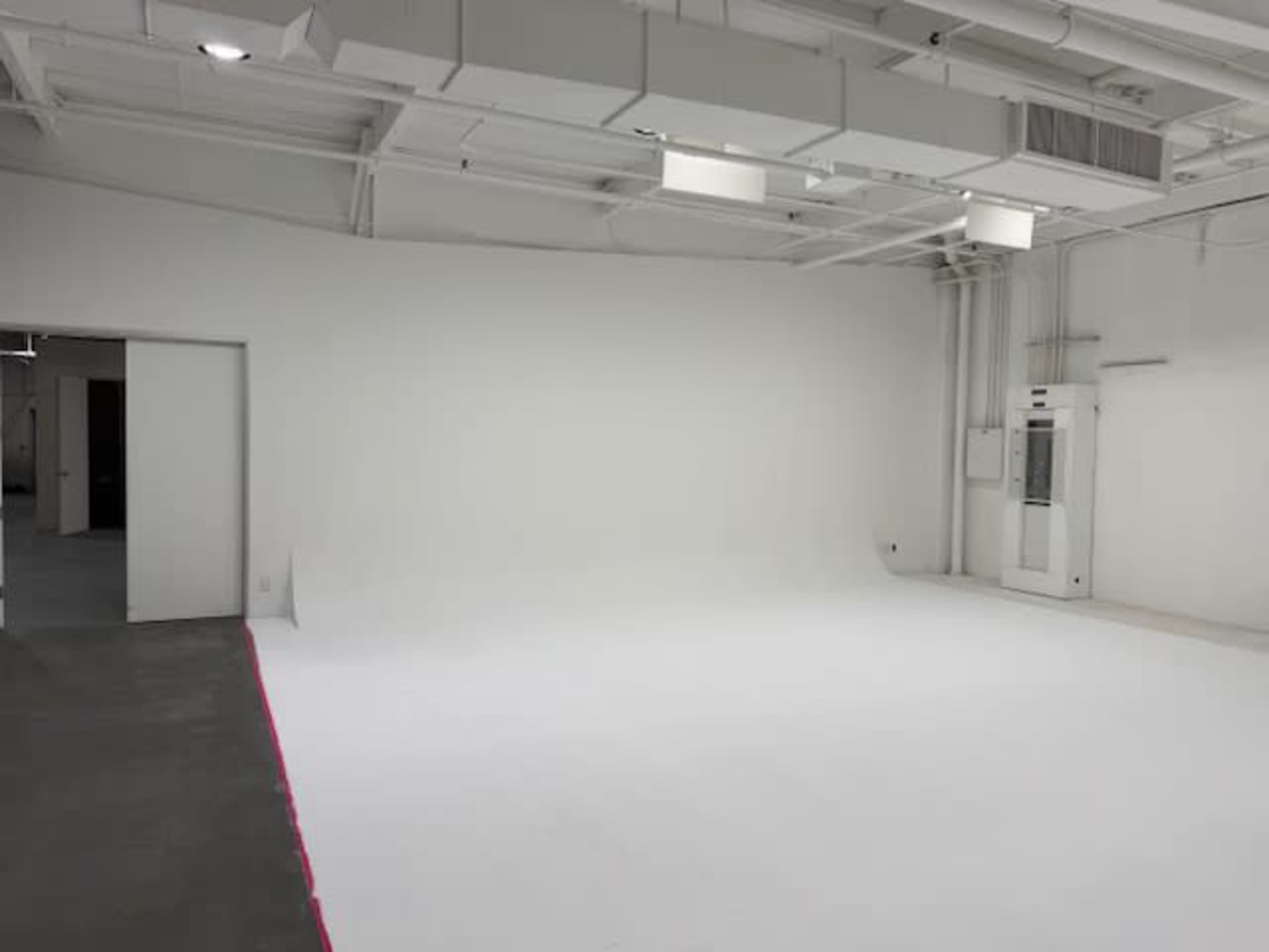 The image shows a spacious, empty room with white walls and a concrete floor, featuring a door on one side and an air conditioning unit on the opposite wall.