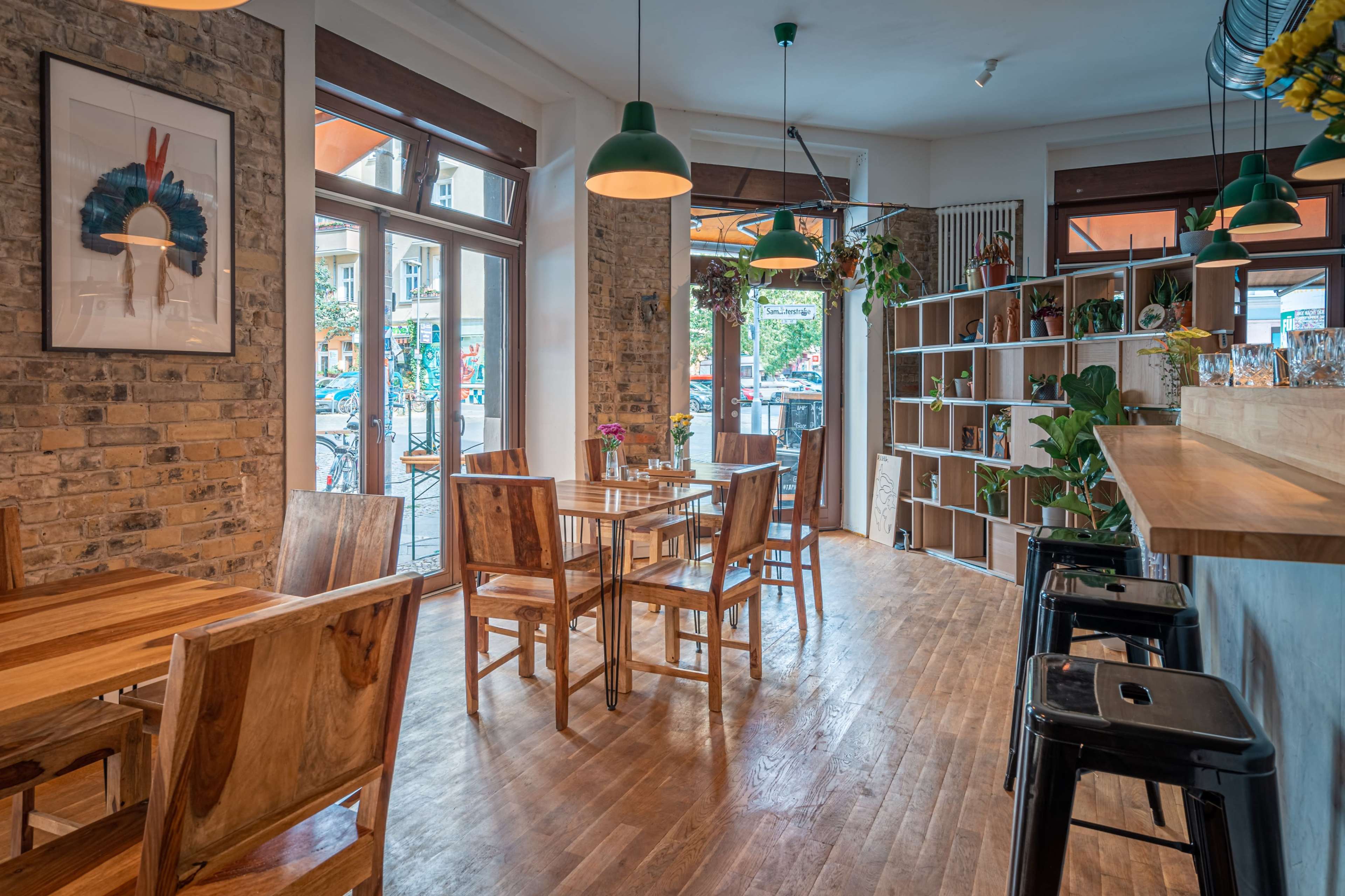 A cozy cafe features wooden tables and chairs, large windows, and plants positioned throughout the space.