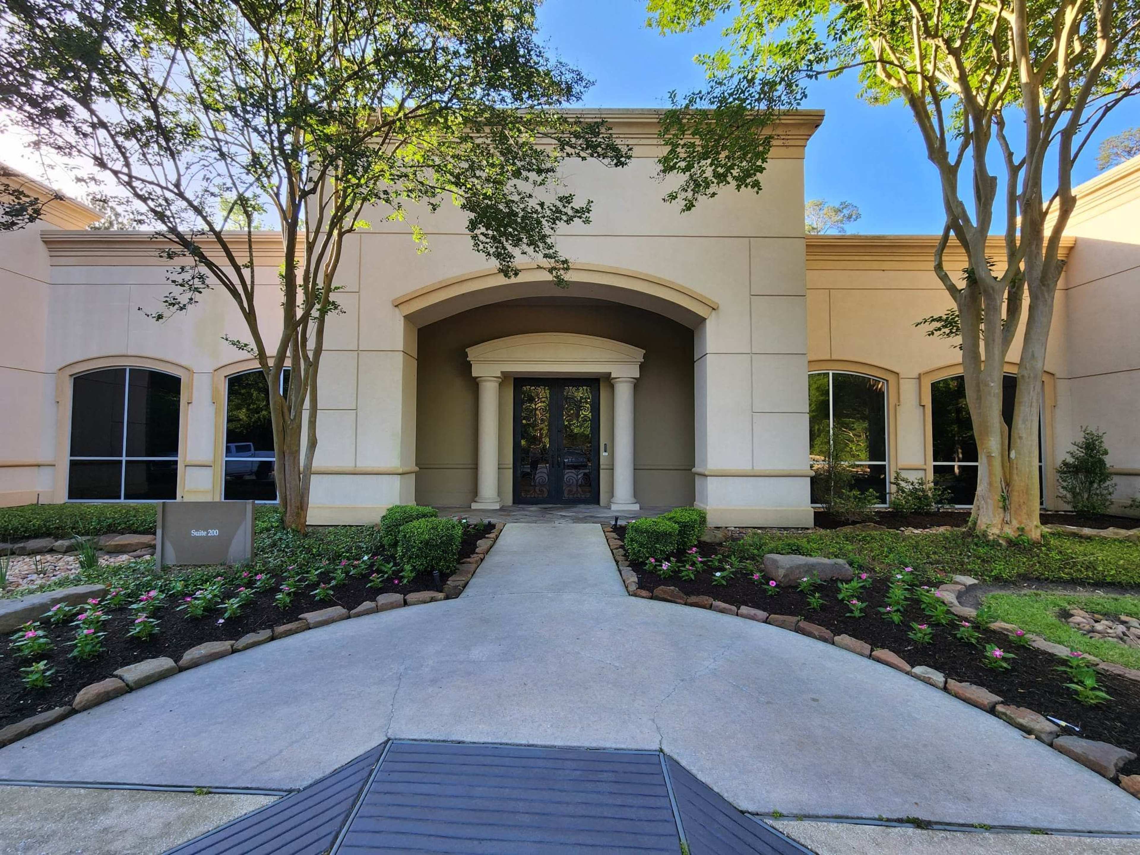 The image shows a modern building entrance surrounded by landscaped gardens with a pathway leading to the doors.