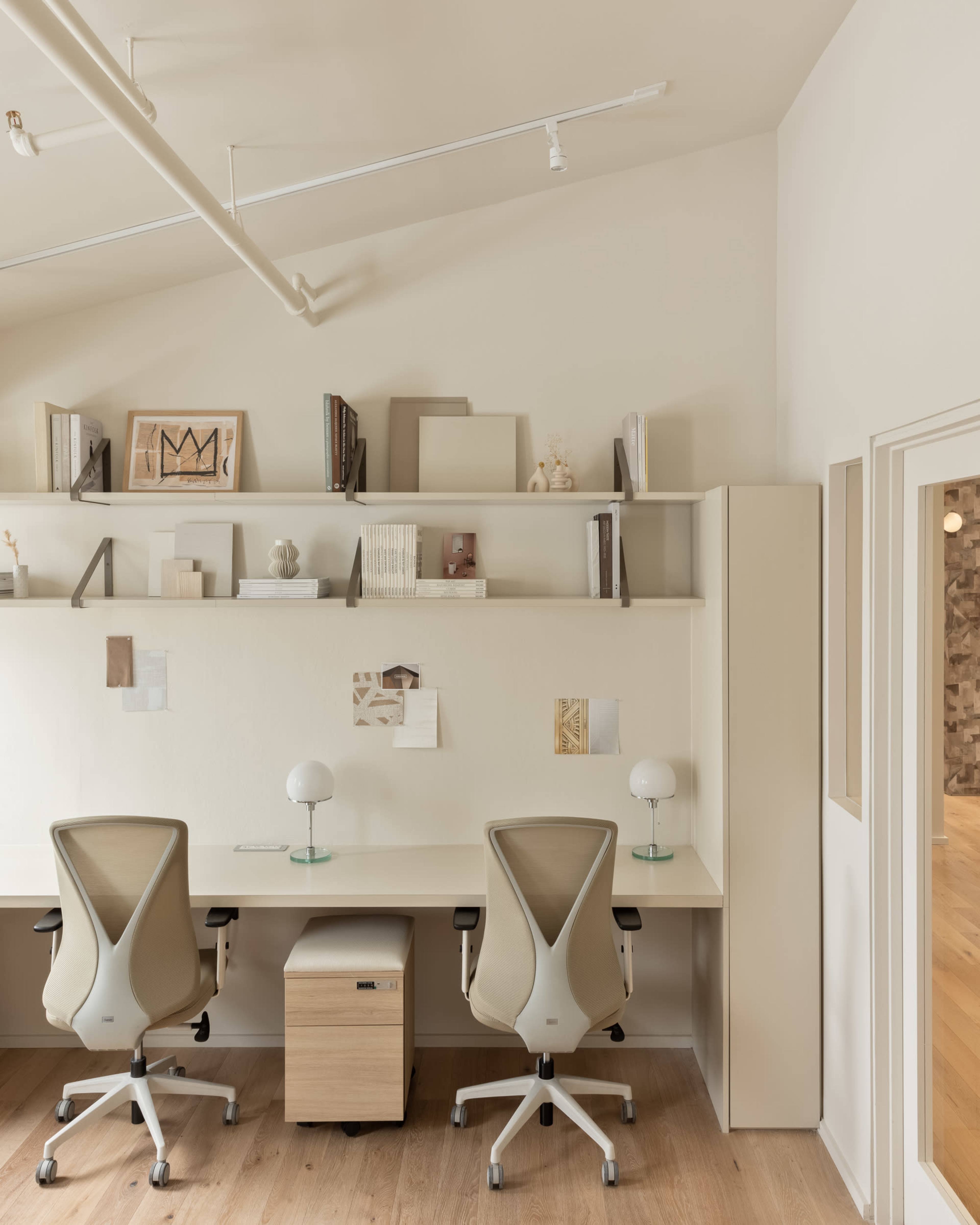 The image shows a minimalist office space with two ergonomic chairs and a wooden desk beneath a shelf displaying various books and decorative items.