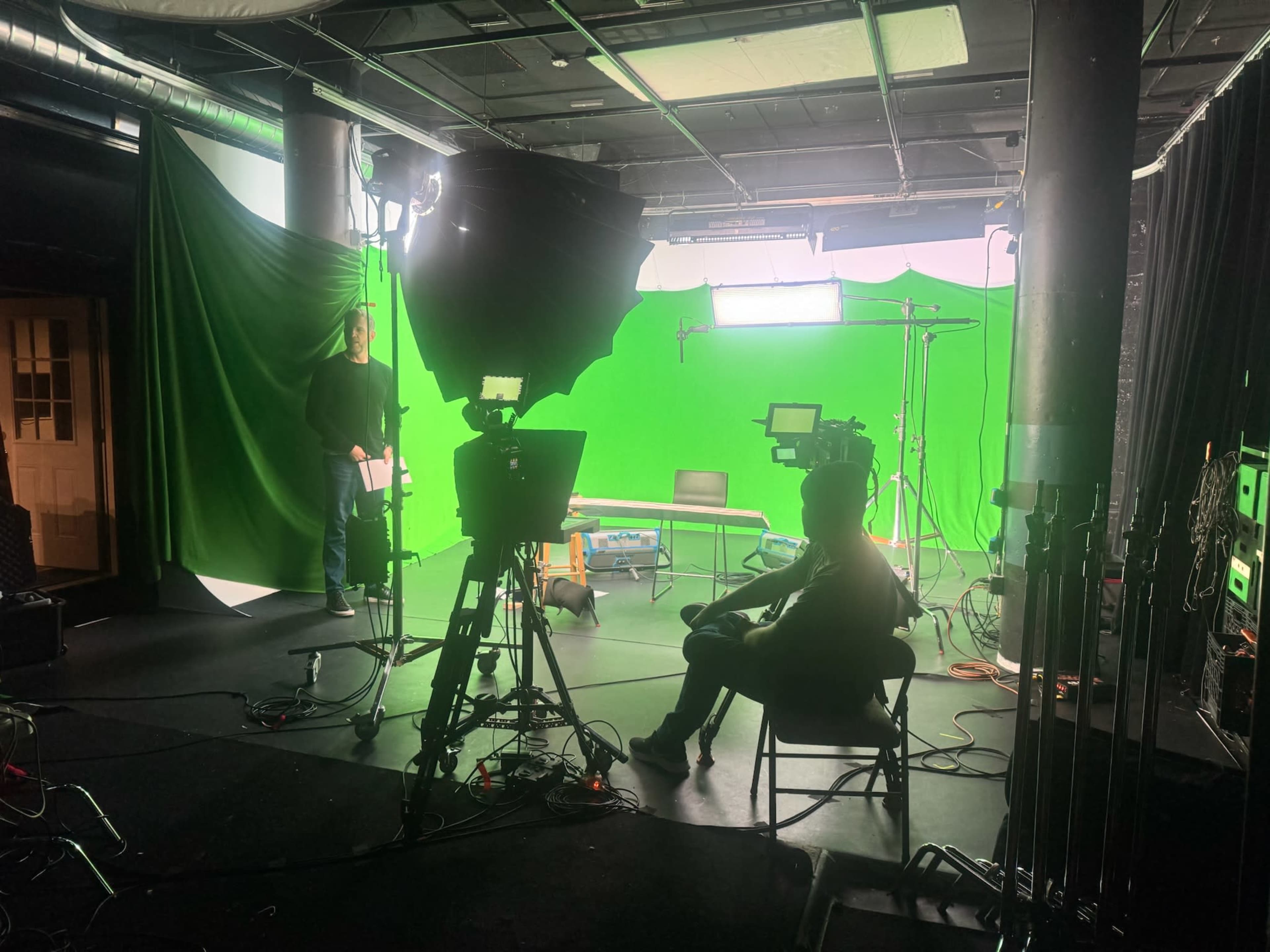 The image shows a film studio with green screens, multiple lights, and two individuals, one seated and the other standing, preparing for a shoot.