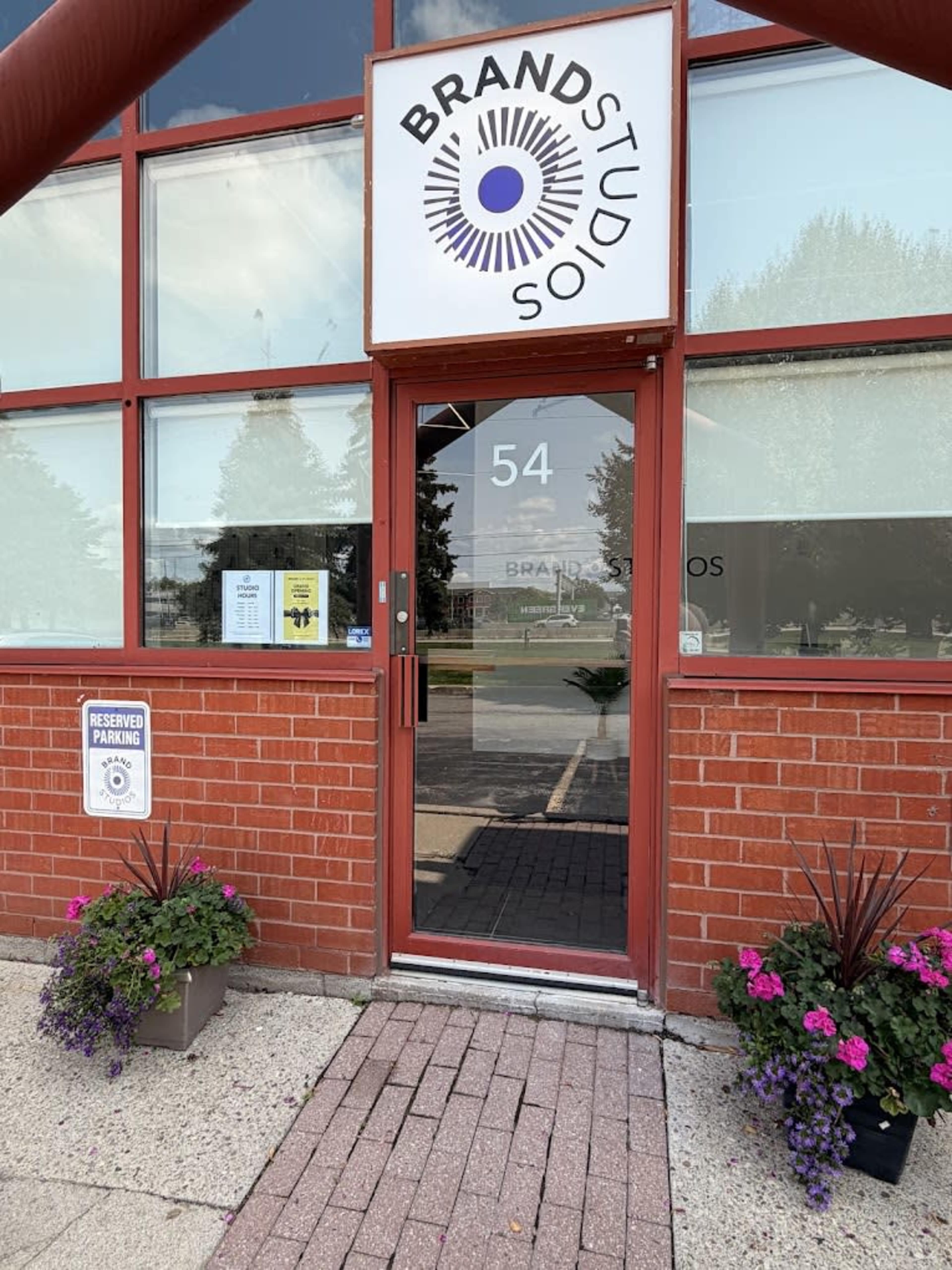 The image shows the entrance of a building labeled "BRAND STUDIO," featuring a glass door and large windows, with flower planters on either side of the entrance.