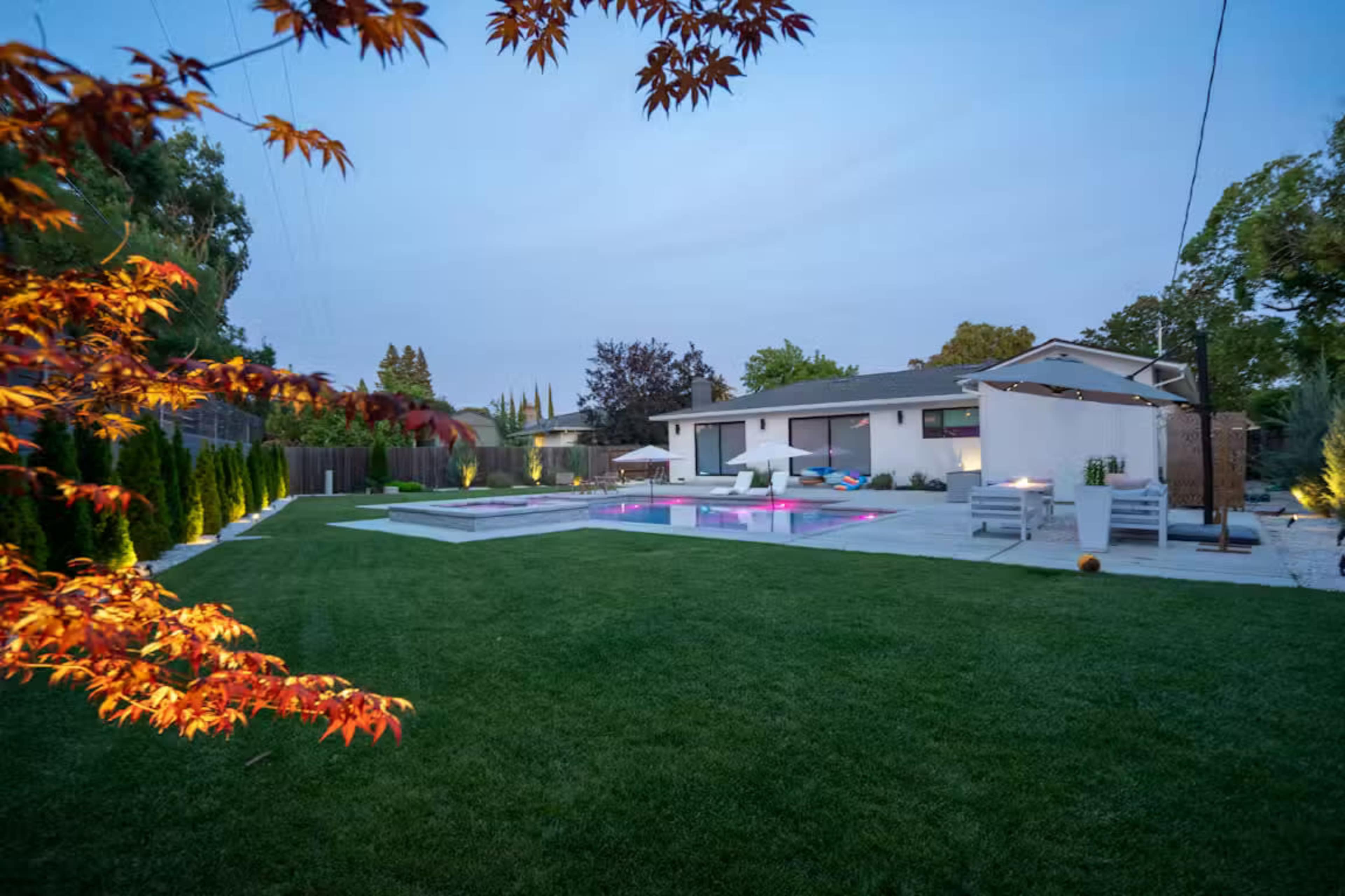 The image shows a modern backyard featuring a swimming pool, landscaped green grass, and a contemporary house with outdoor seating.