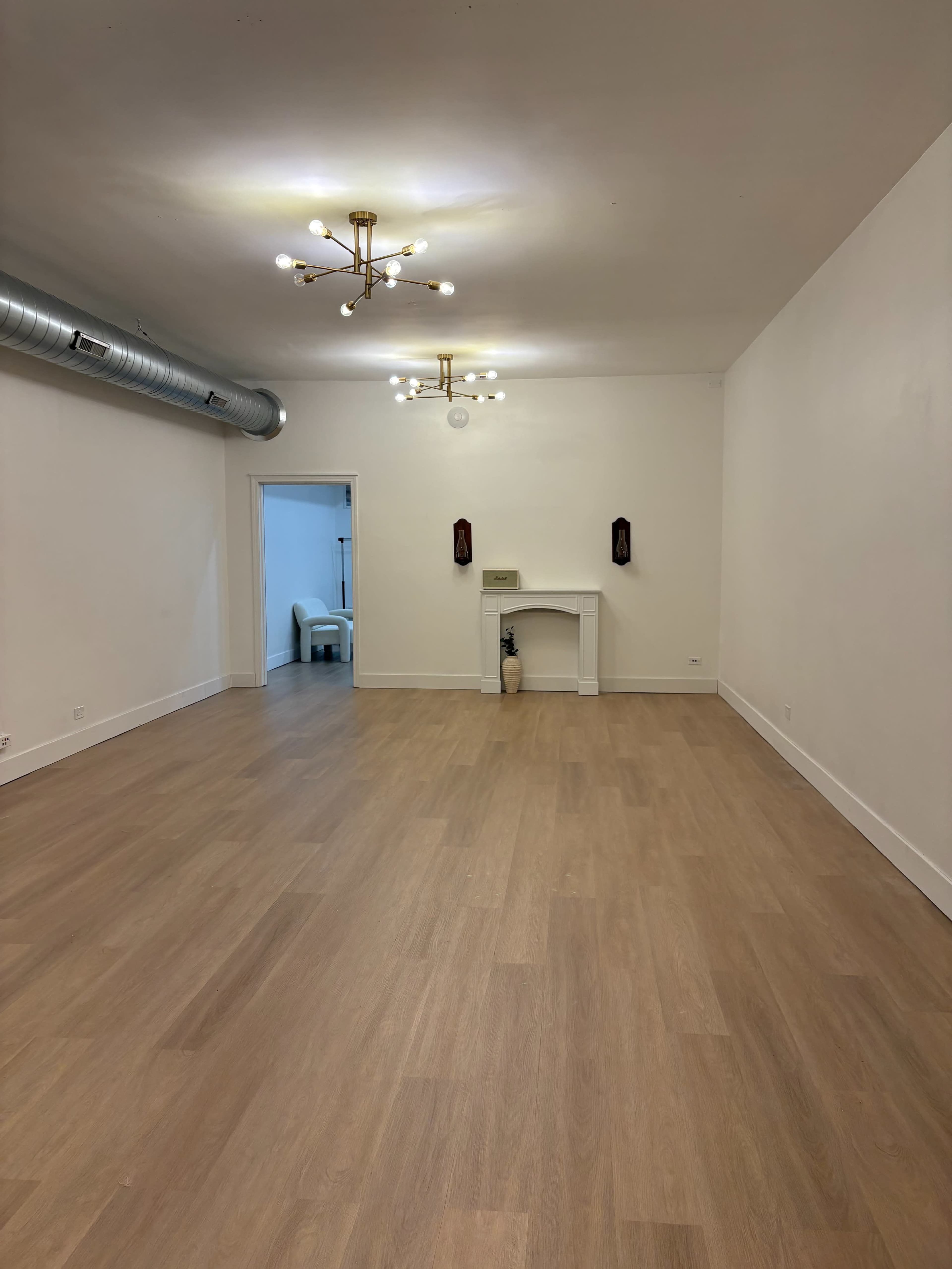The image shows a spacious room with light wood flooring, white walls, a decorative fireplace, and lighting fixtures on the ceiling.