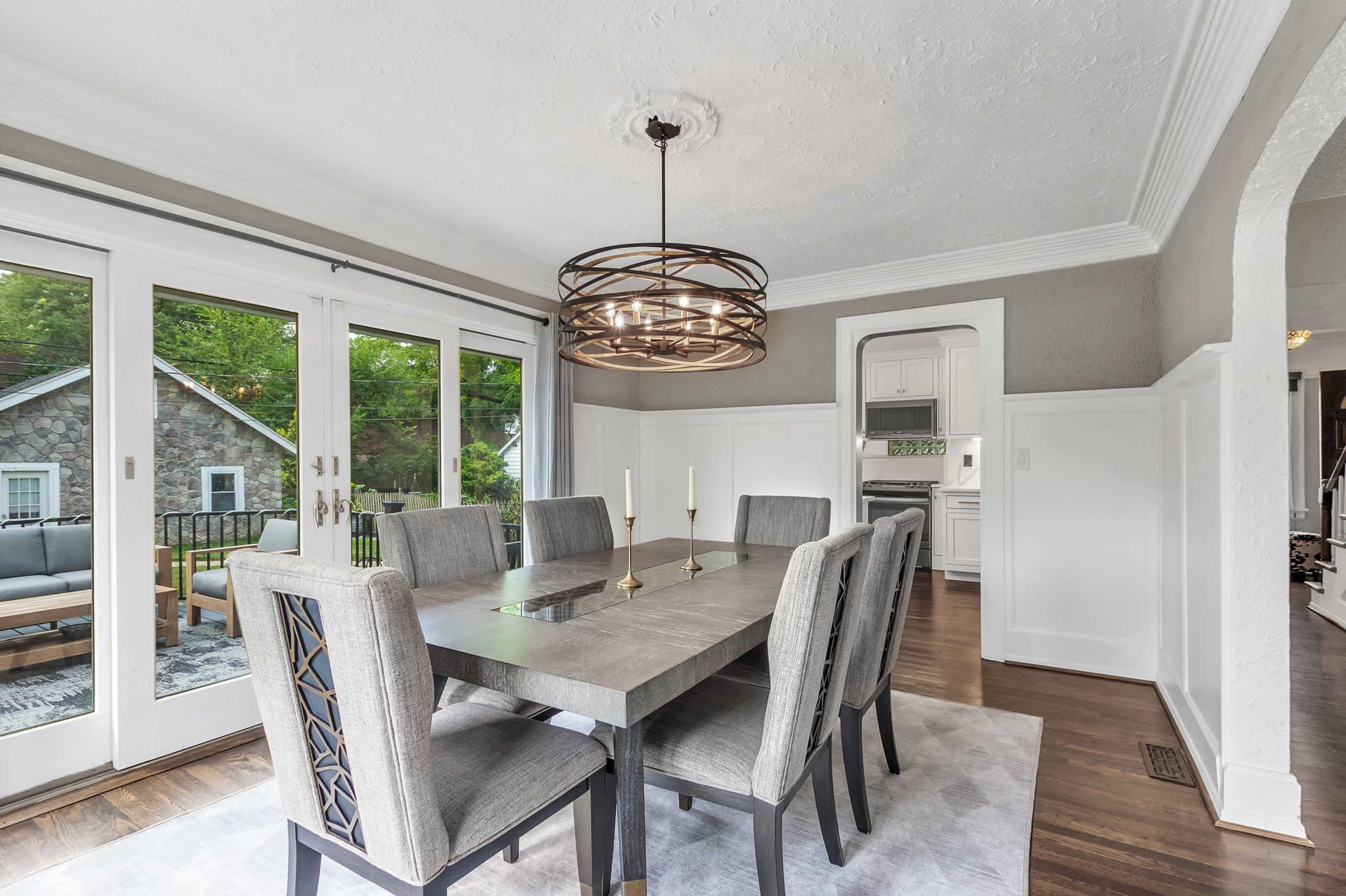 A dining area with a rectangular table set for six, featuring a modern light fixture above, and large glass doors that open to an outdoor space.