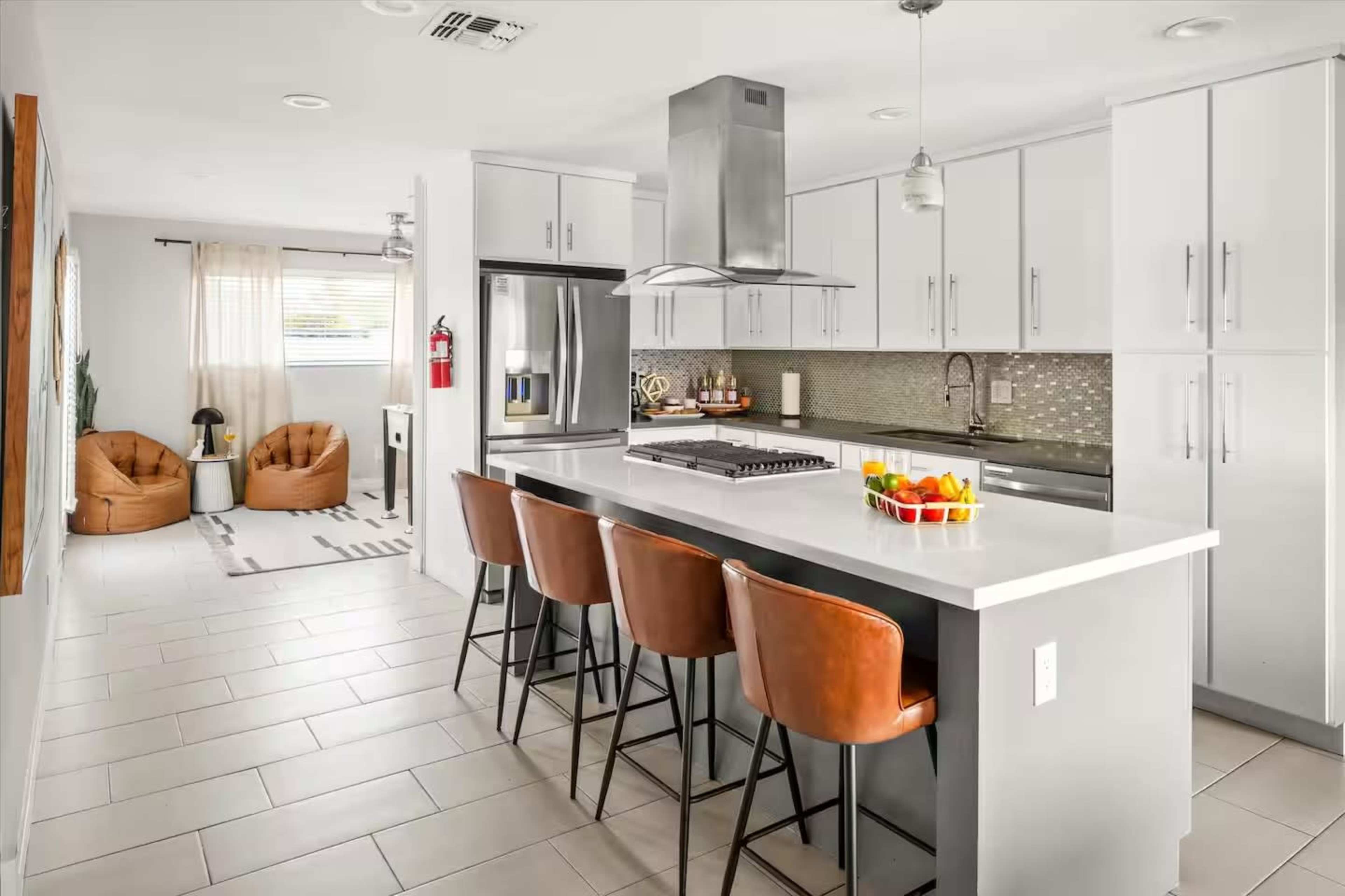 The image shows a modern kitchen with a central island featuring four bar stools, stainless steel appliances, and a living area visible in the background.