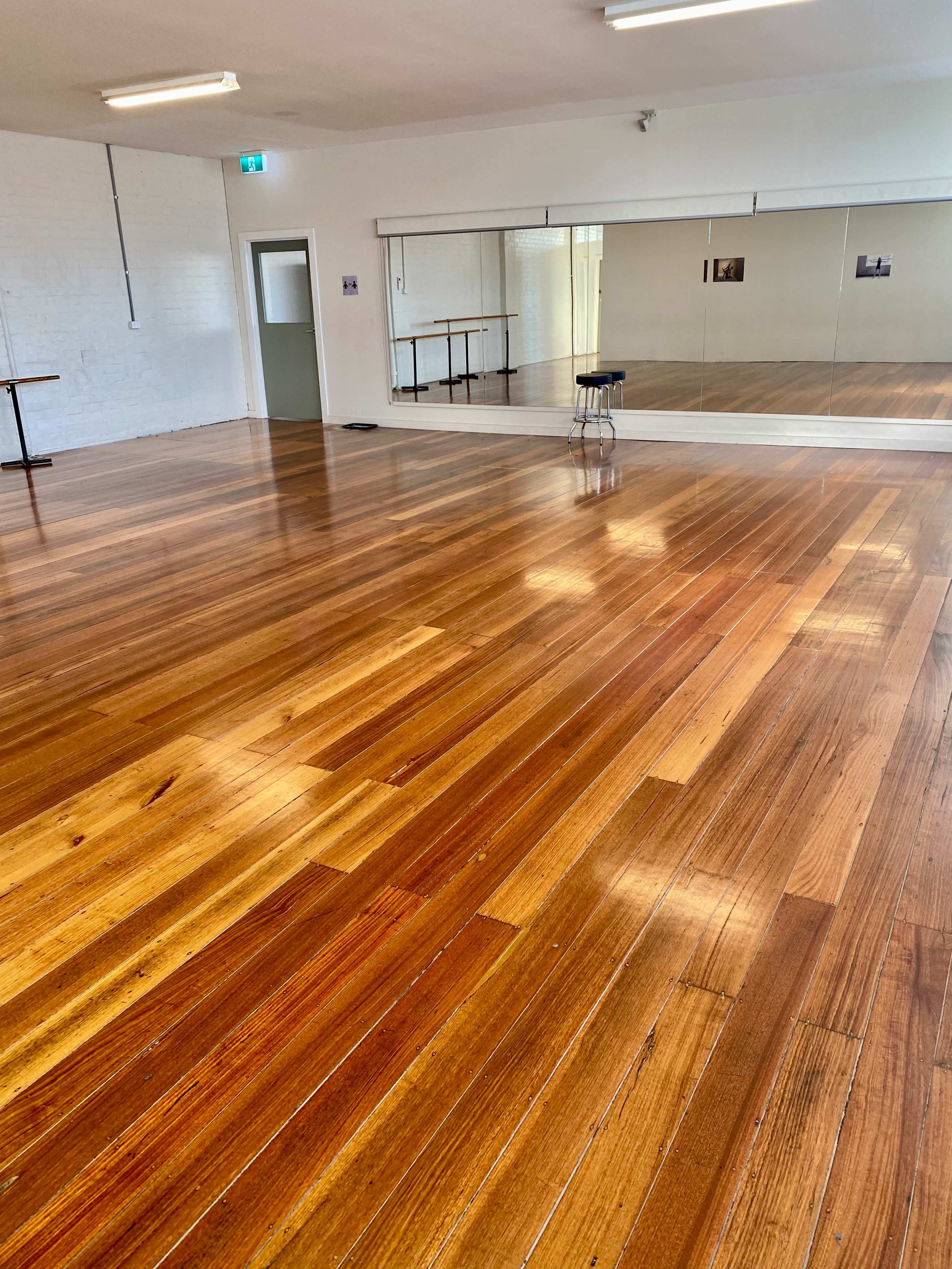 The image shows a spacious room with polished wooden floors, bright overhead lights, and a large mirror along one wall.