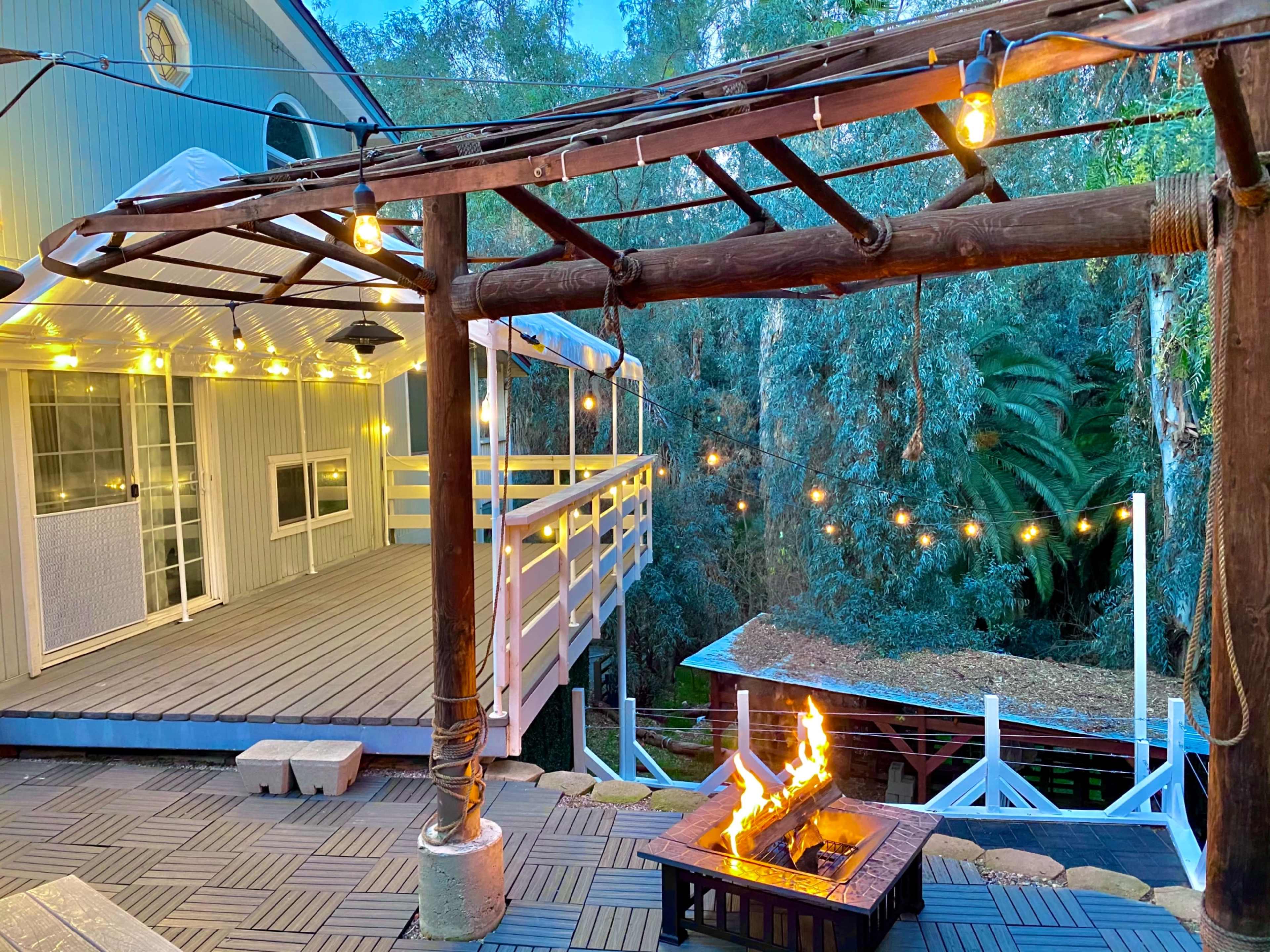 A wooden pergola adorned with string lights frames a fire pit, positioned near a deck surrounded by trees.