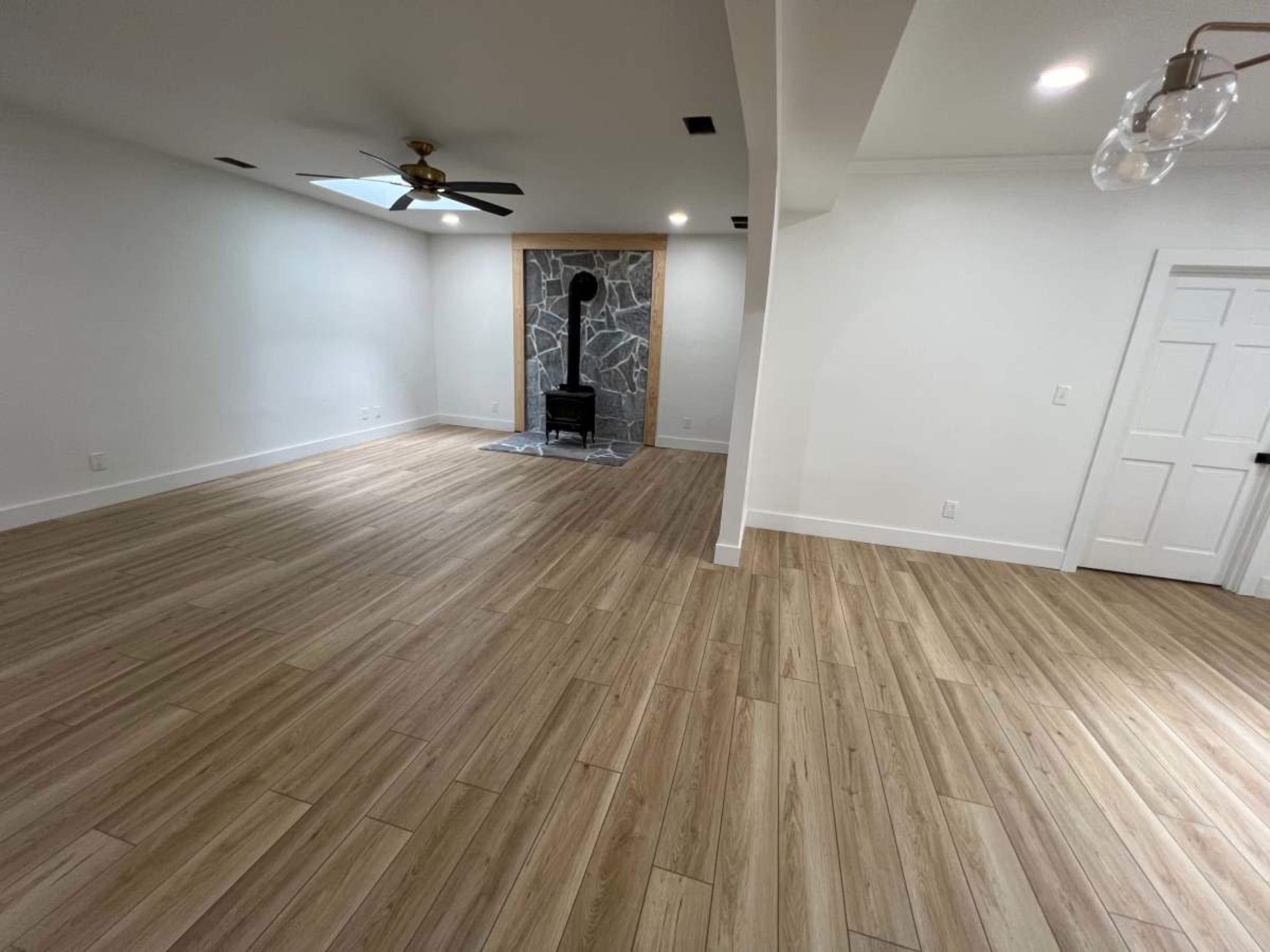 A spacious, empty room with light wood flooring, a ceiling fan, and a stone fireplace at one end.