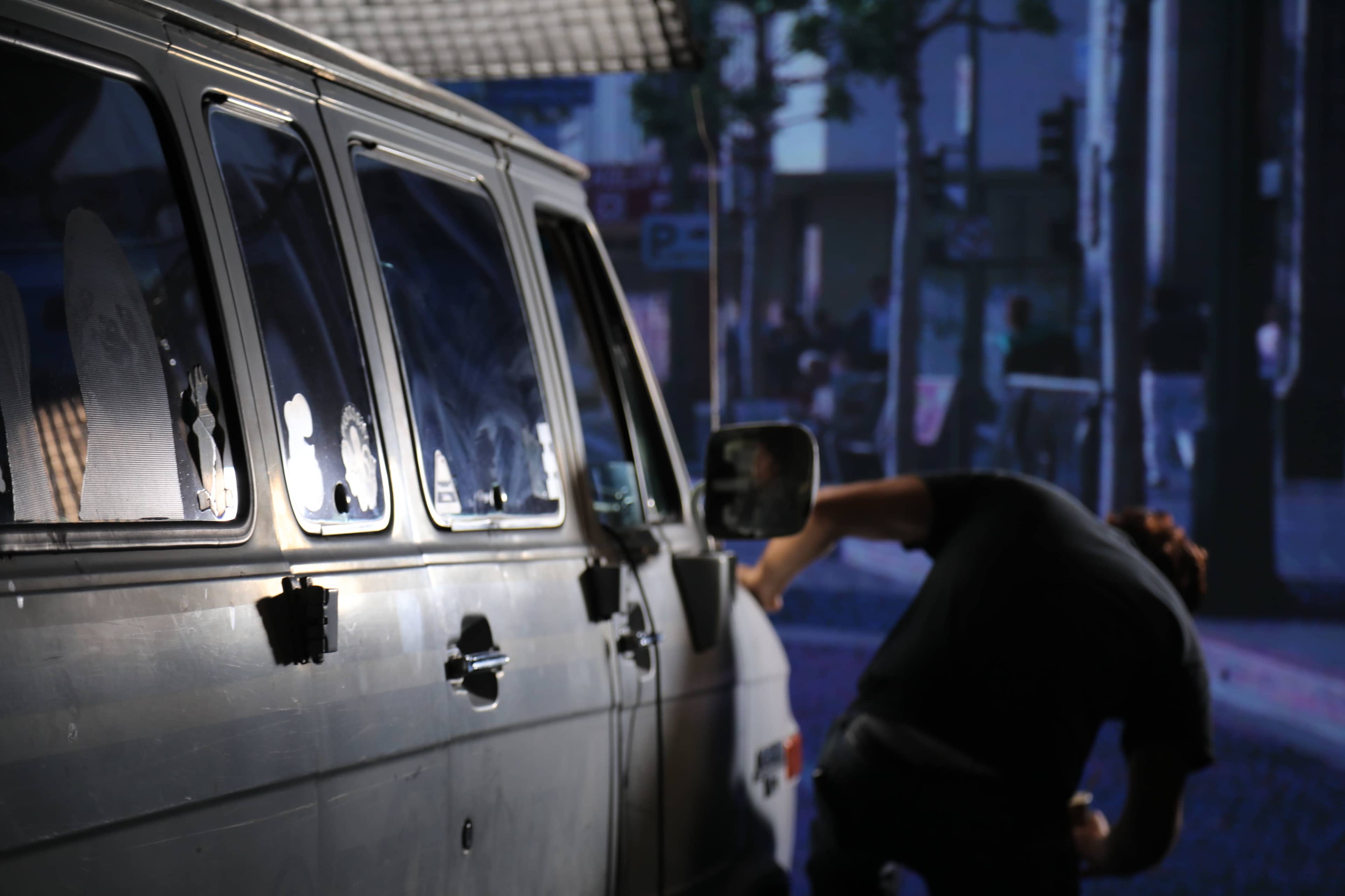 A person leans over to inspect the side of a silver van parked along a city street.