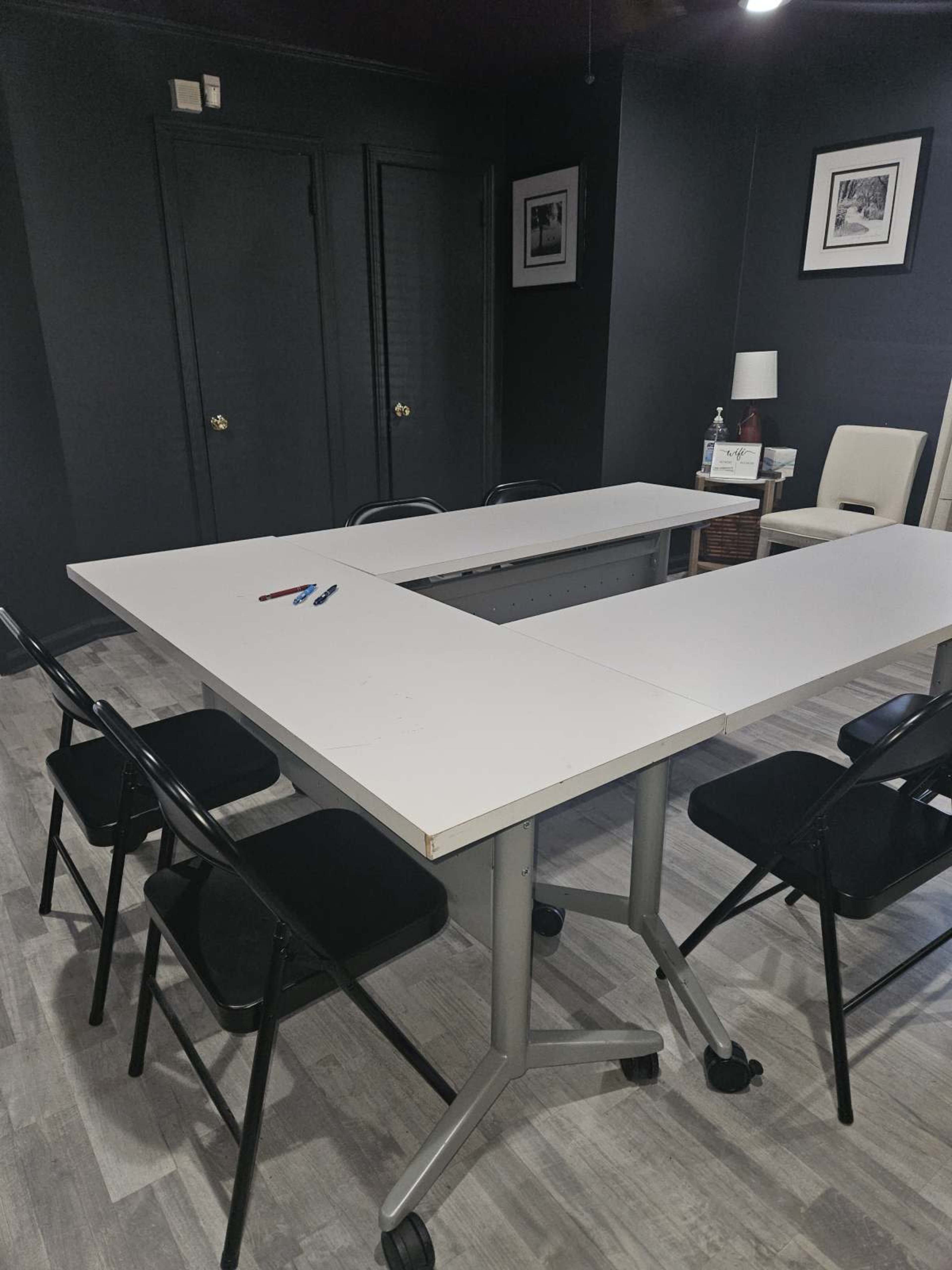 A rectangular meeting table with six black chairs is set up in a dimly lit room with dark walls and framed pictures.