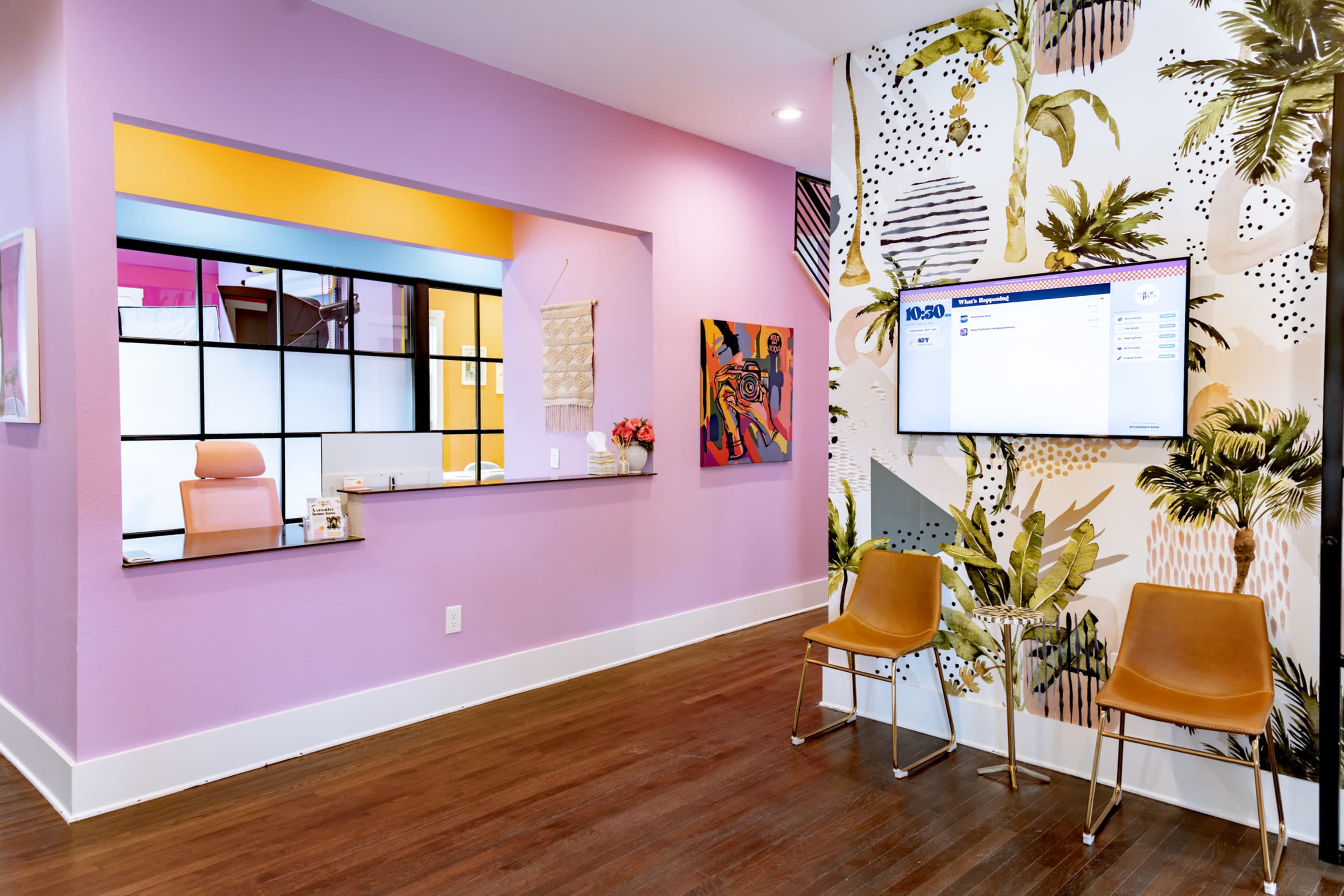 The image shows a brightly colored waiting area with a pink and white tropical-themed wall, a seating area with two chairs, and a reception window in the background.