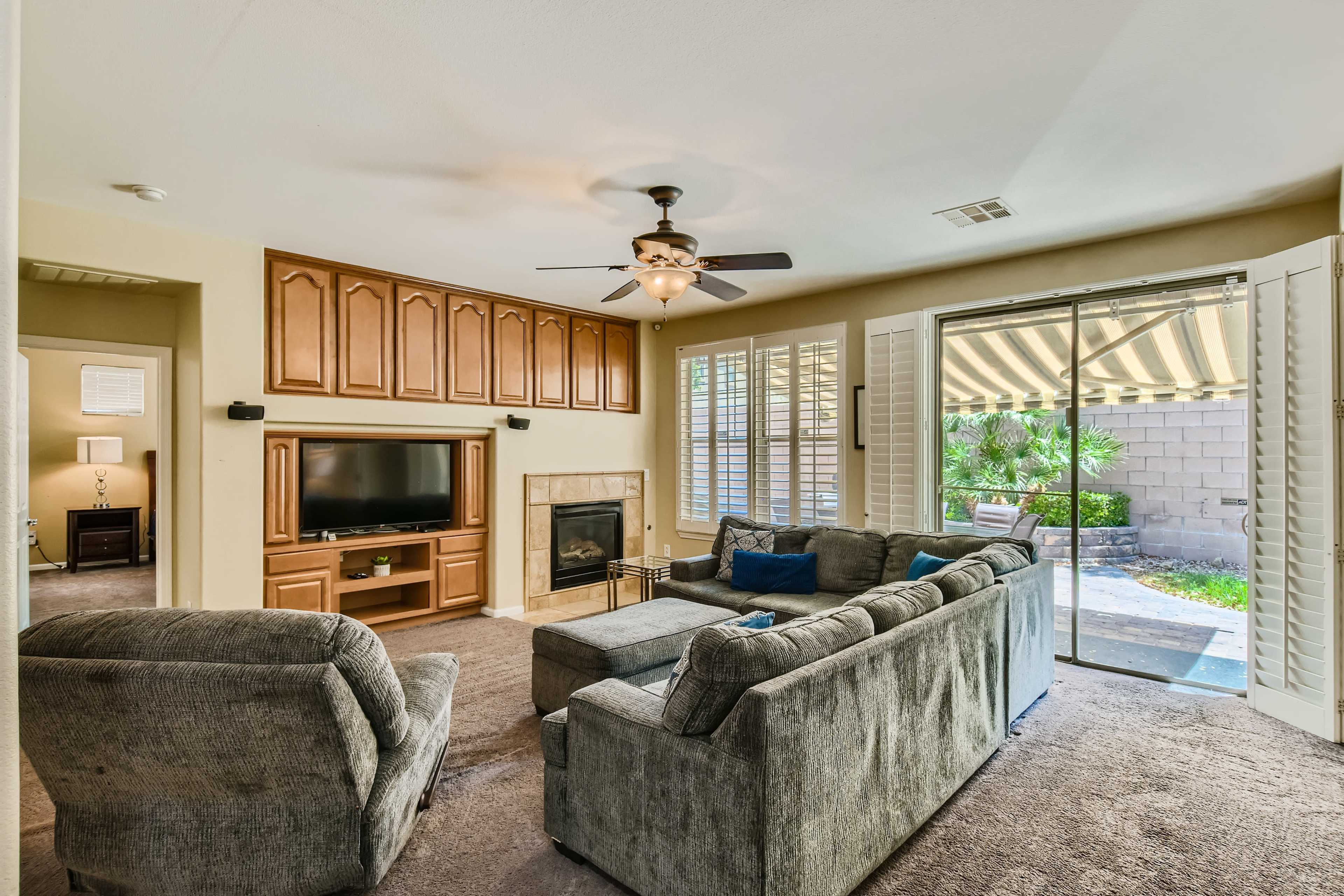 The living room features a ceiling fan, a wall-mounted television, a fireplace, and large windows leading to an outdoor patio.