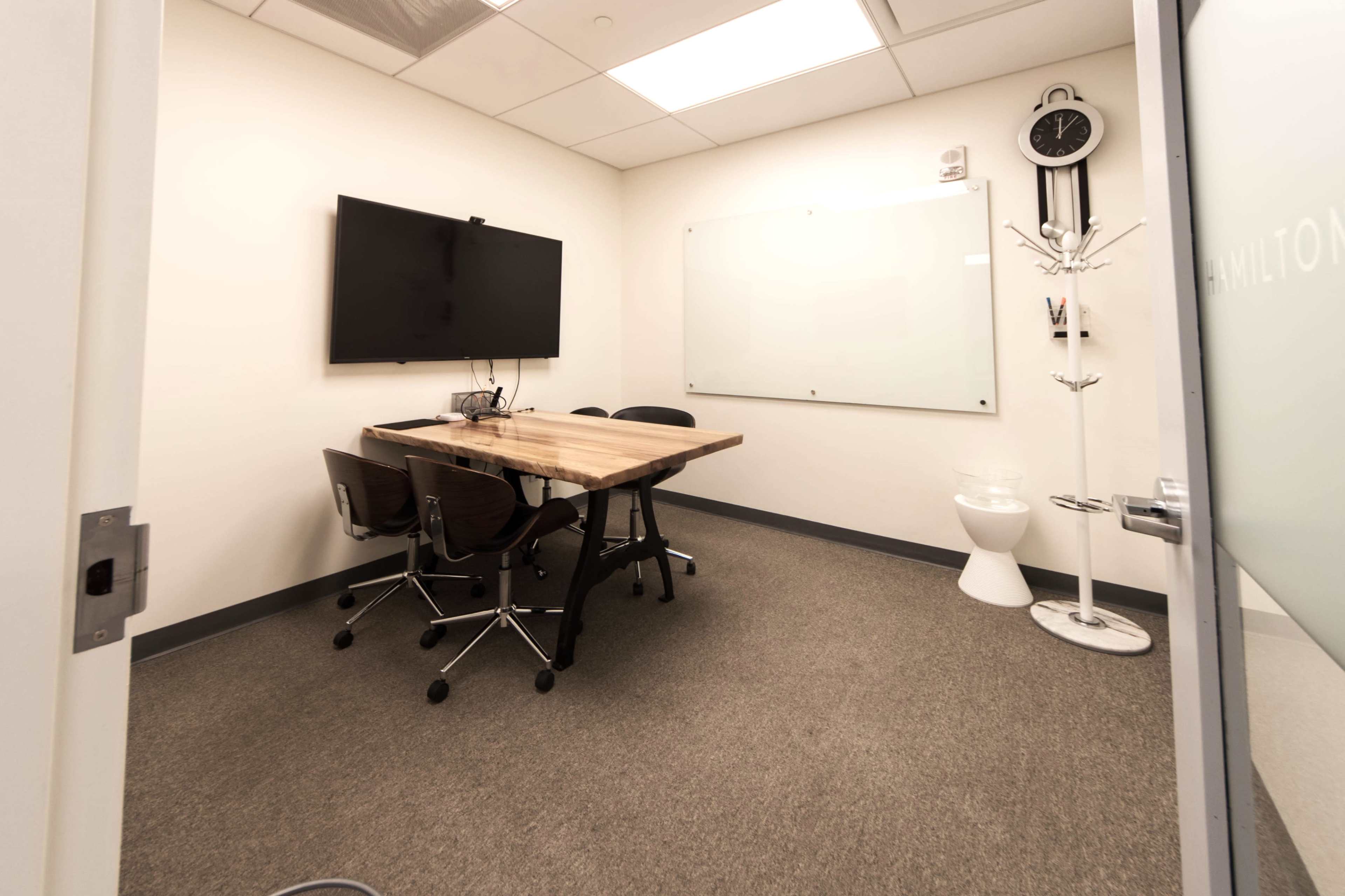 The image shows a small conference room featuring a wooden table with four chairs, a large screen mounted on the wall, and a whiteboard.