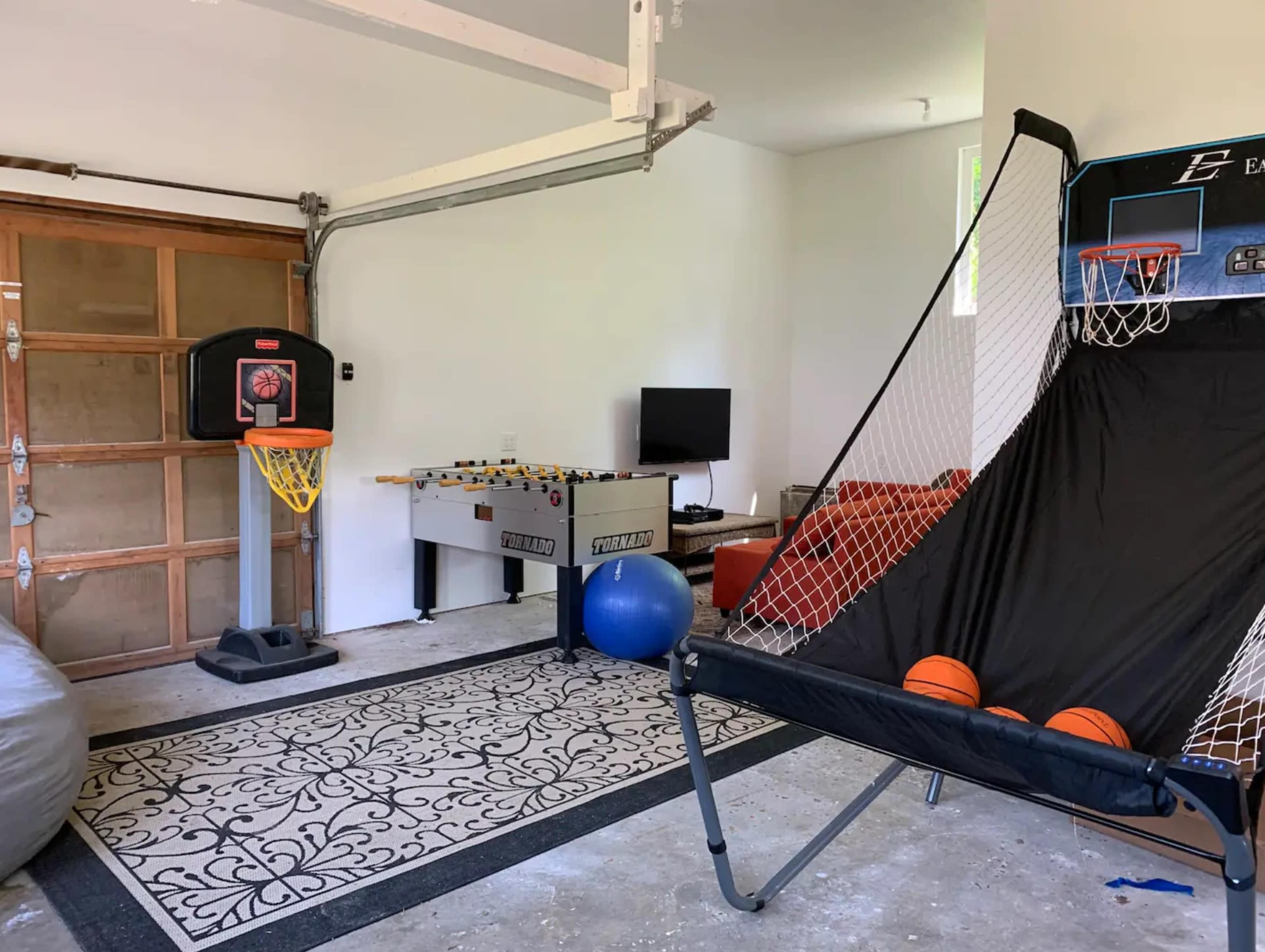 A garage space equipped with a basketball hoop, a foosball table, a television, a couch, and a blue exercise ball on a patterned rug.